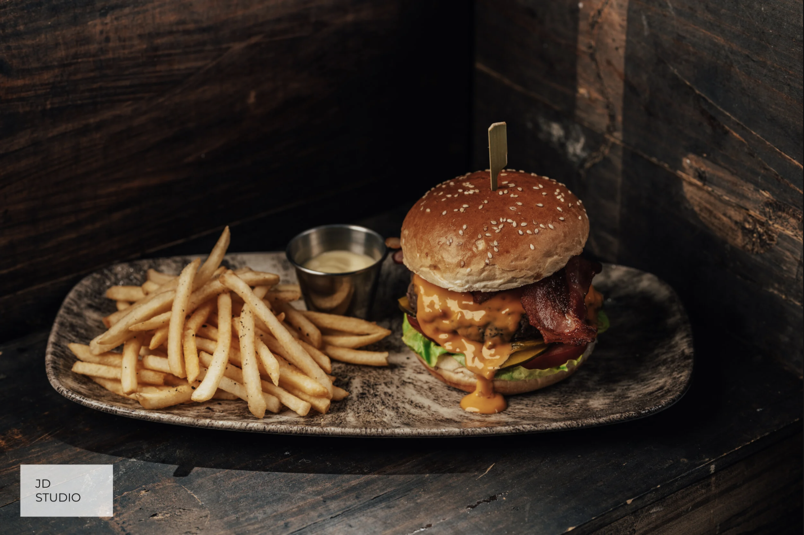 A large burger with bacon, cheese, lettuce, tomato, and a bun, served with French fries and dipping sauce on a gray platter