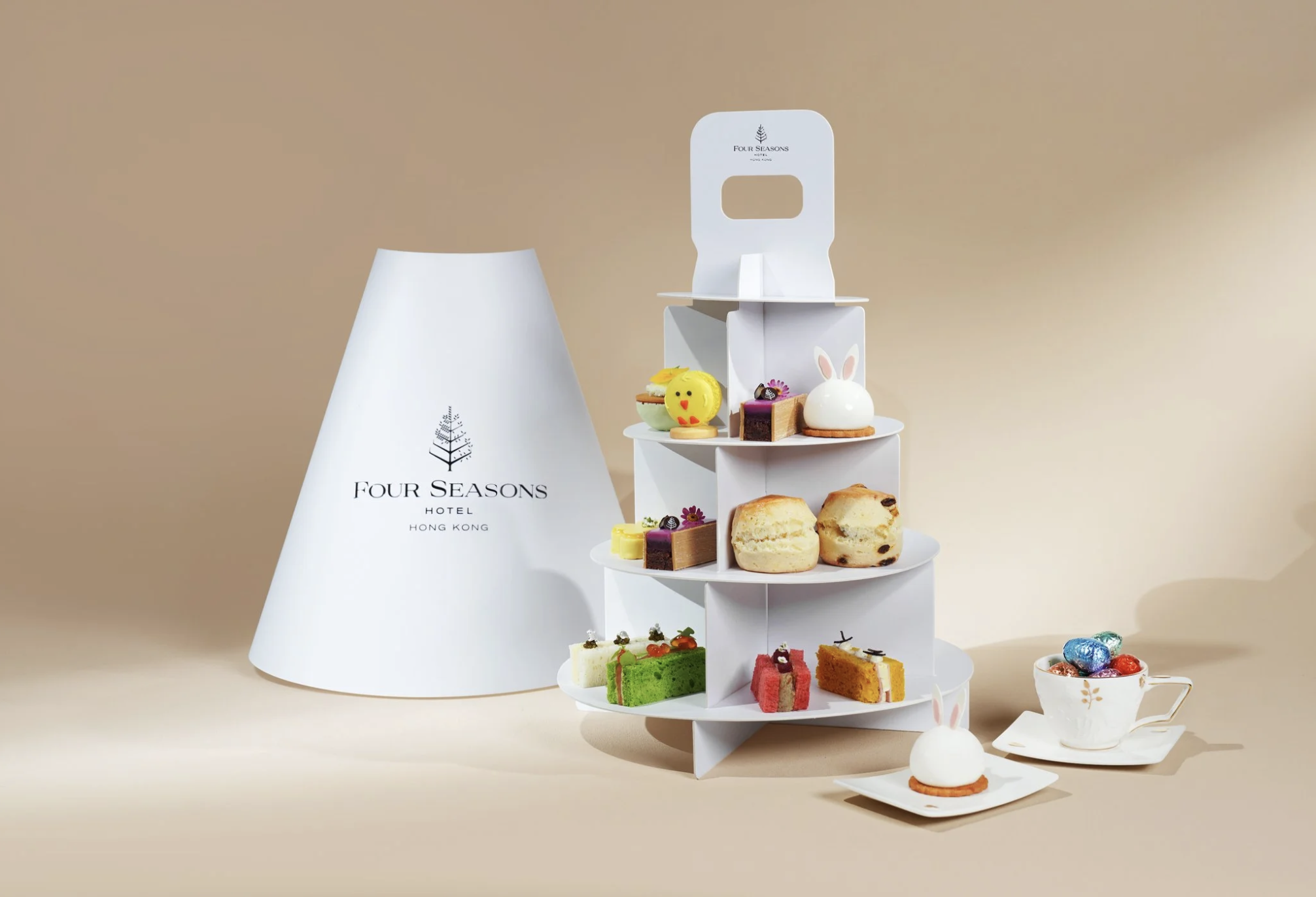 A white tiered dessert stand with various decorated cakes and pastries, including a bunny-shaped cake, a yellow duck-shaped cake, a chocolate cake slice, and fruit-topped pastries, next to a white cup of colorful chocolates and a white cone-shaped bo