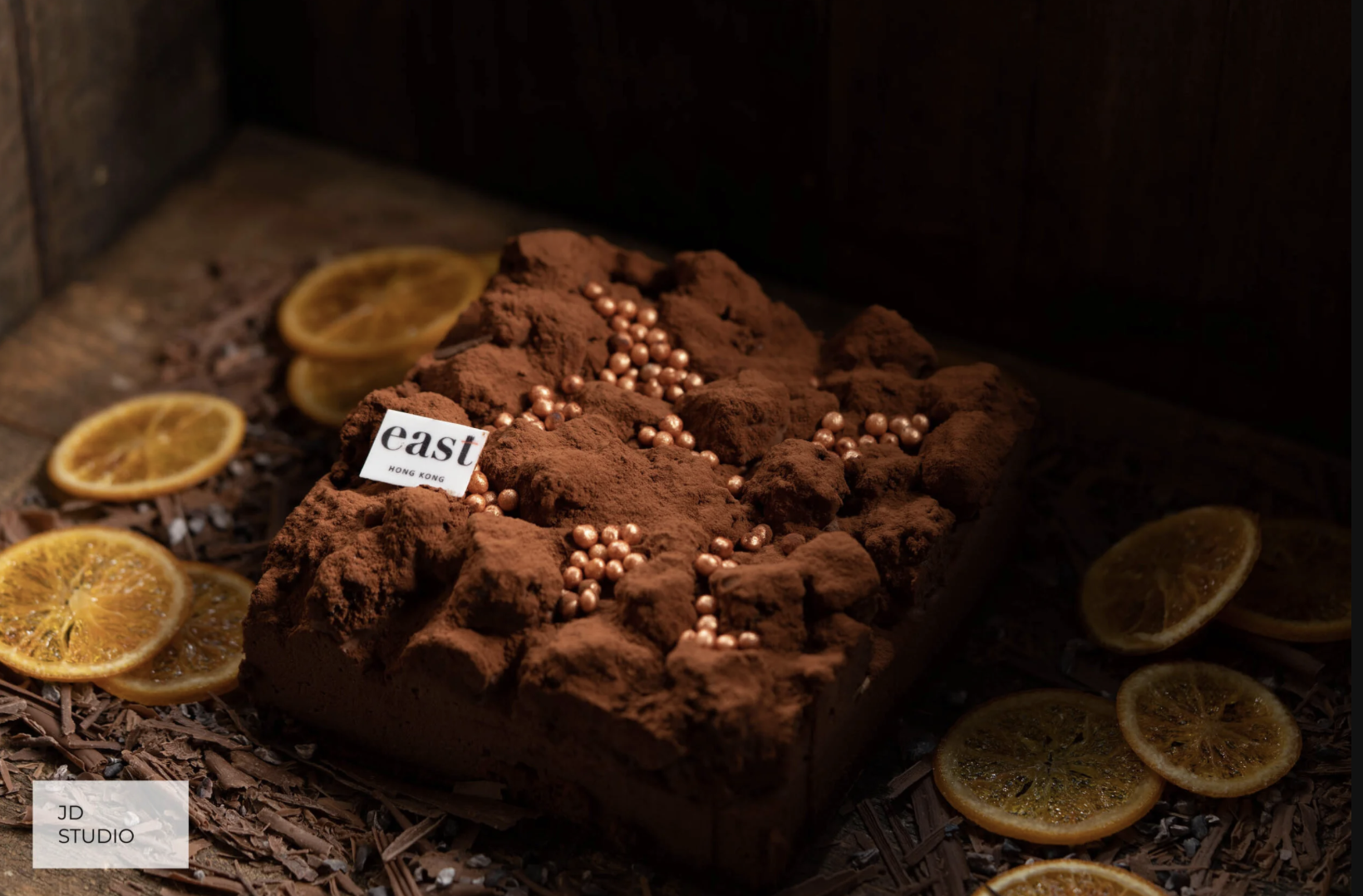 A rectangular chocolate dessert topped with edible pearls and cocoa powder, garnished with sliced dried oranges around it, and a small tag that says 'east HONG KONG'.