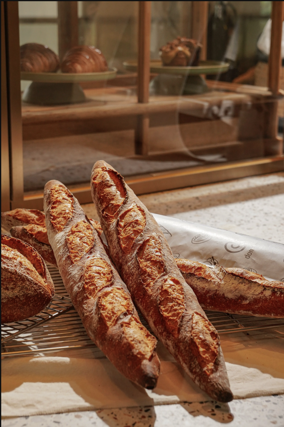 Fresh baguettes on a cooling rack in a bakery, with a display case containing pastries in the background.