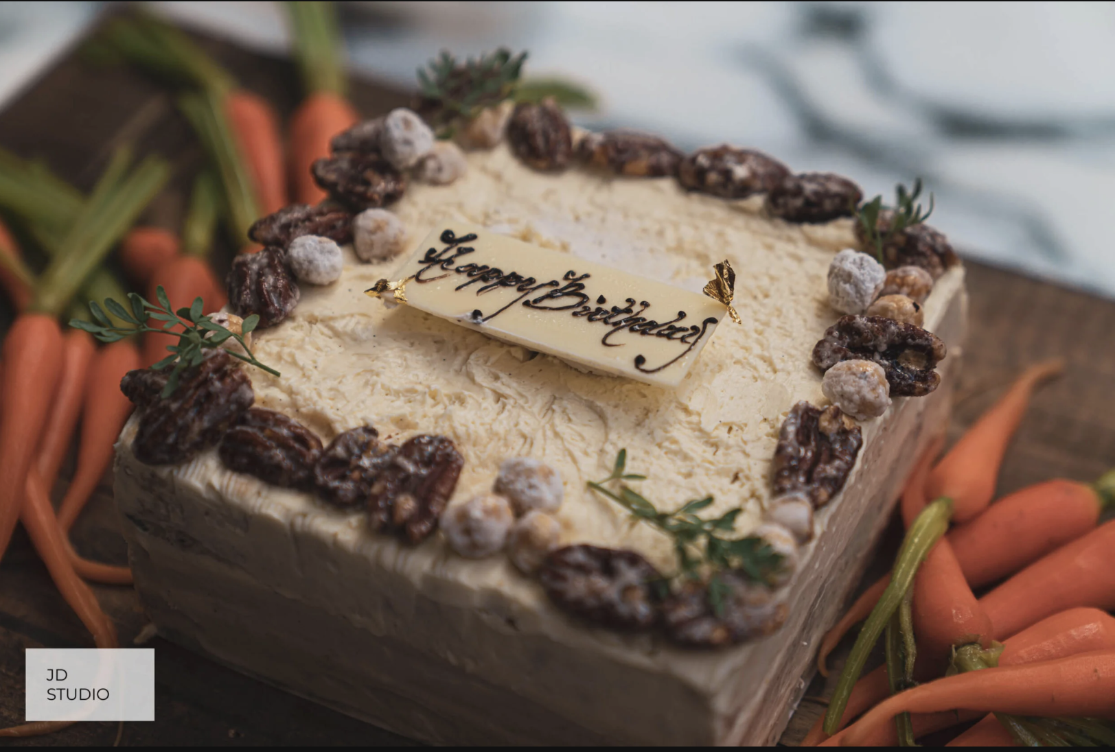 Square birthday cake with cream frosting, decorated with pecans, small white powdered sugar toppings, green herbs, and a white chocolate plaque that says 'Happy Birthday' in black script. Surrounding the cake are fresh carrots with green tops.