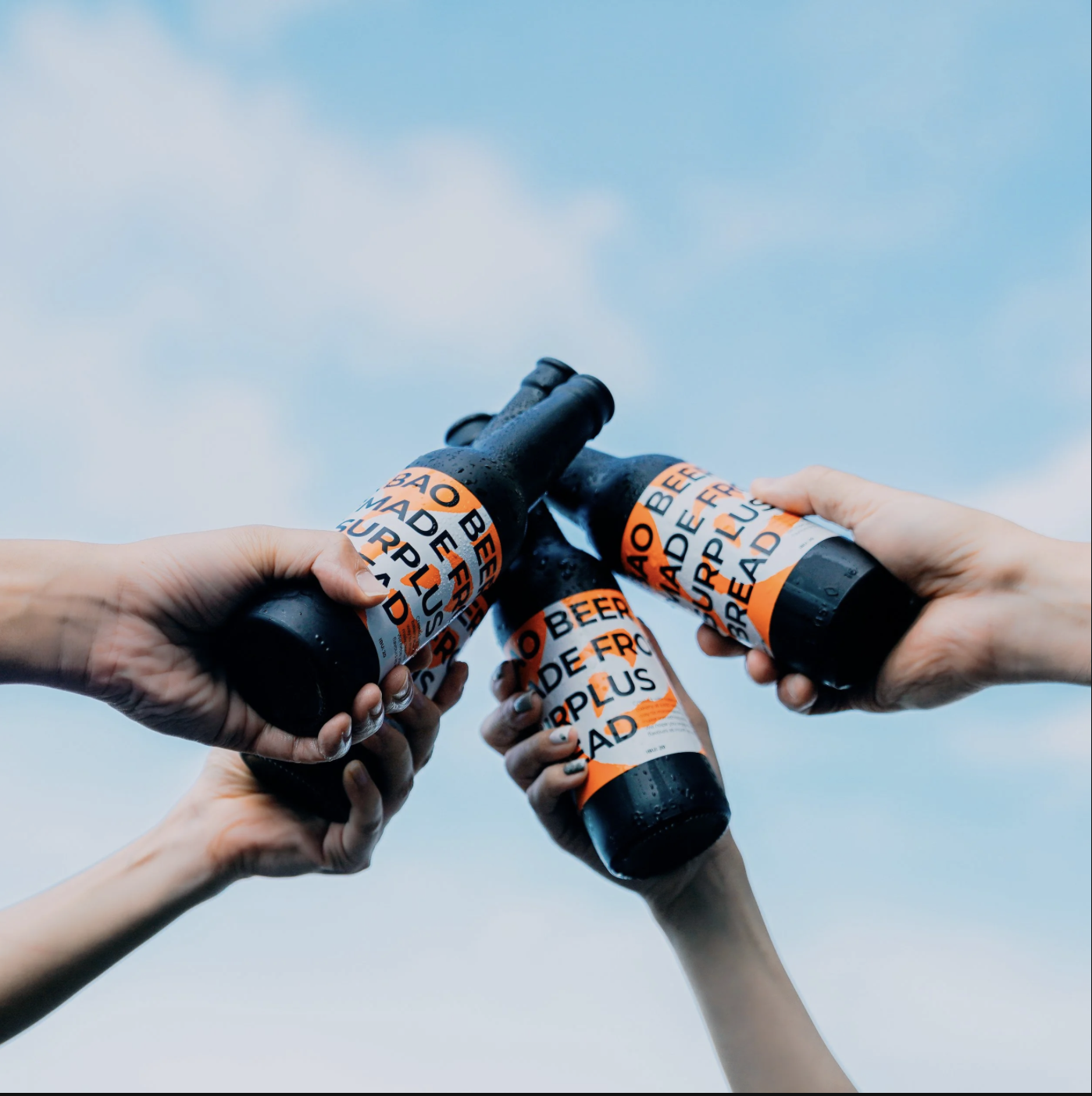 Four hands holding beer bottles with the labels wrapped in orange and white caution tape that reads 'Beef Made Free Plus' against a cloudy sky background.