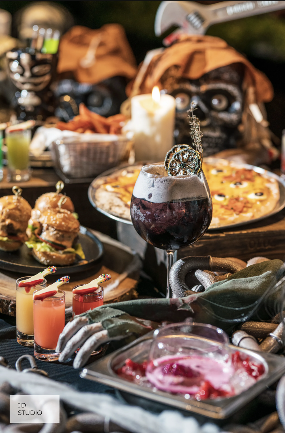 A spooky Halloween-themed spread of food and drinks, featuring a skeleton hand jewelry, a glass of red wine with dried citrus garnish, a pizza, mini sandwiches, colorful drinks with sprinkles, and a lit candle among Halloween decorations including a 