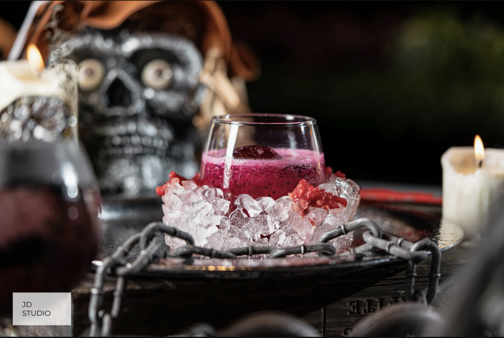 A glass of dark pink beverage with ice and strawberries in a bowl surrounded by ice, candles, and Halloween decorations including a black skull with eyes, featuring a Halloween or Day of the Dead theme.
