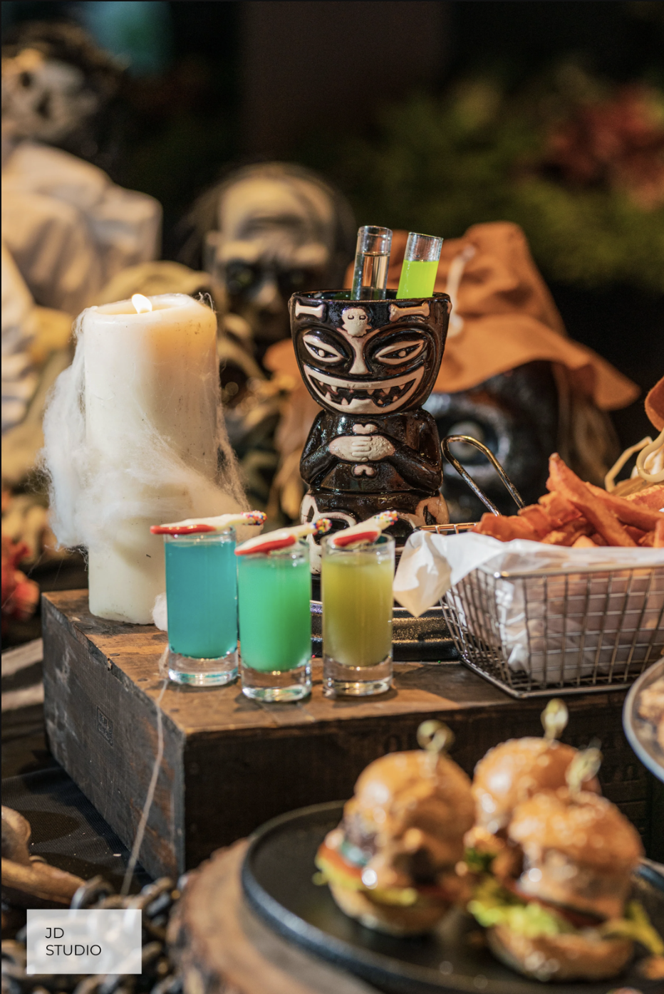 Halloween-themed table with a carved black ceramic container holding test tubes, colorful drinks with umbrella stir sticks, a large melting candle, fried food in a basket, and ghost-shaped burgers with toothpicks, set against a backdrop of Halloween 