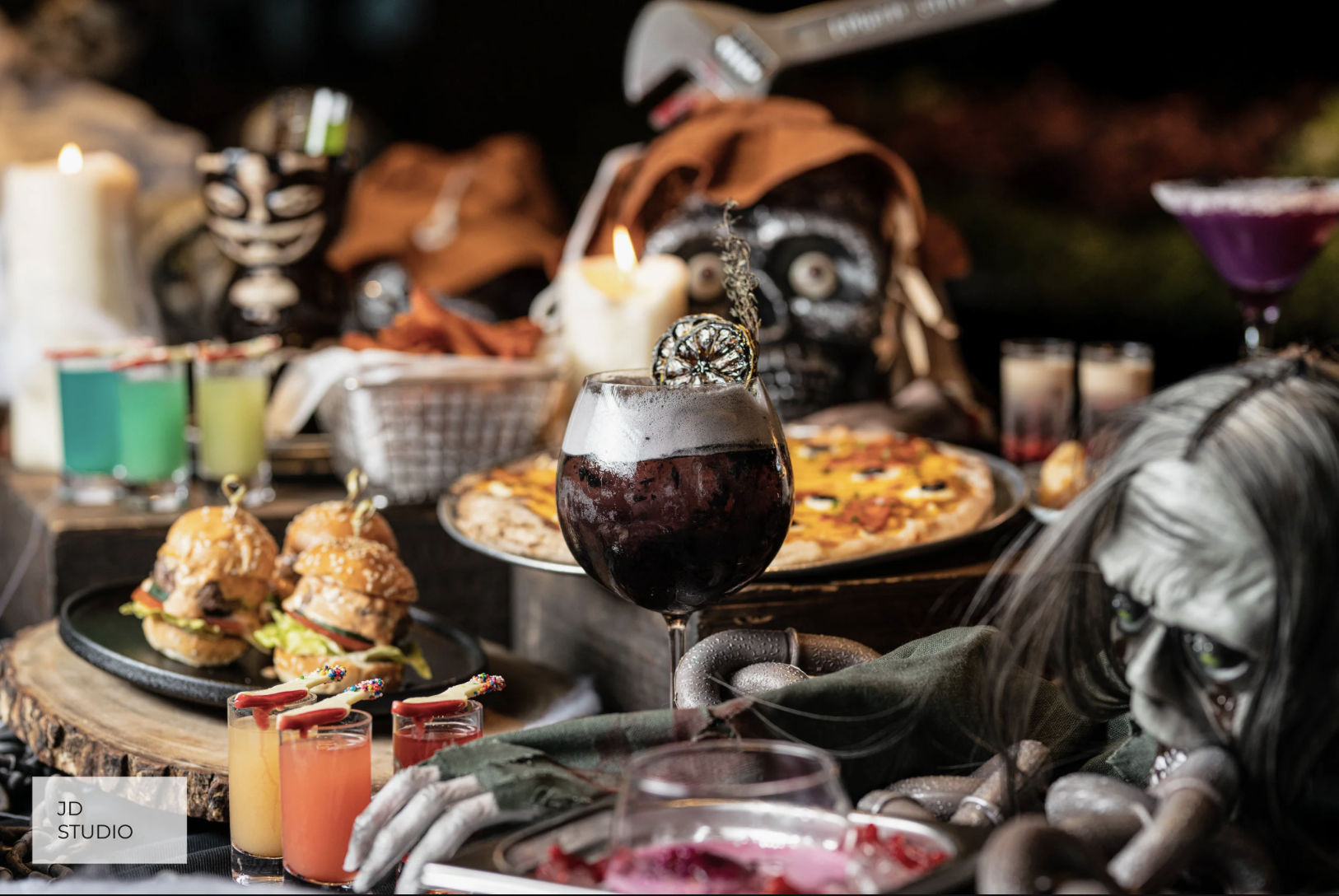 Halloween themed table with food and decorations, including a dark cocktail garnished with a slice of dried citrus, mini burgers, pizza, colorful drinks, candles, and decorative skulls and skeletons.