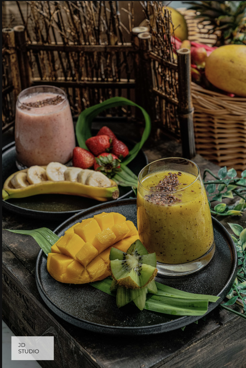 A black tray with a yellow smoothie topped with chia seeds, a sliced mango, and a sliced kiwi in the foreground. In the background, there is a pink smoothie garnished with cacao nibs, strawberries, sliced bananas, and a basket of fruits, including ap