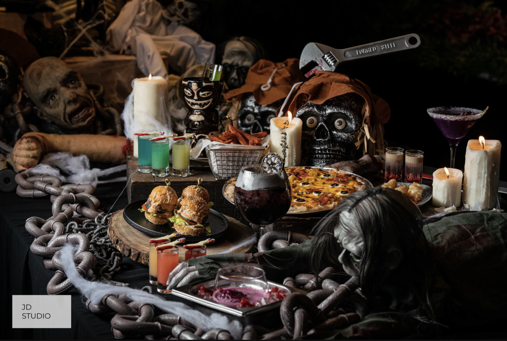 A spooky Halloween-themed table with skulls, skeletons, candles, drinks, pizza, sliders, and Halloween decorations including chains and cobwebs.