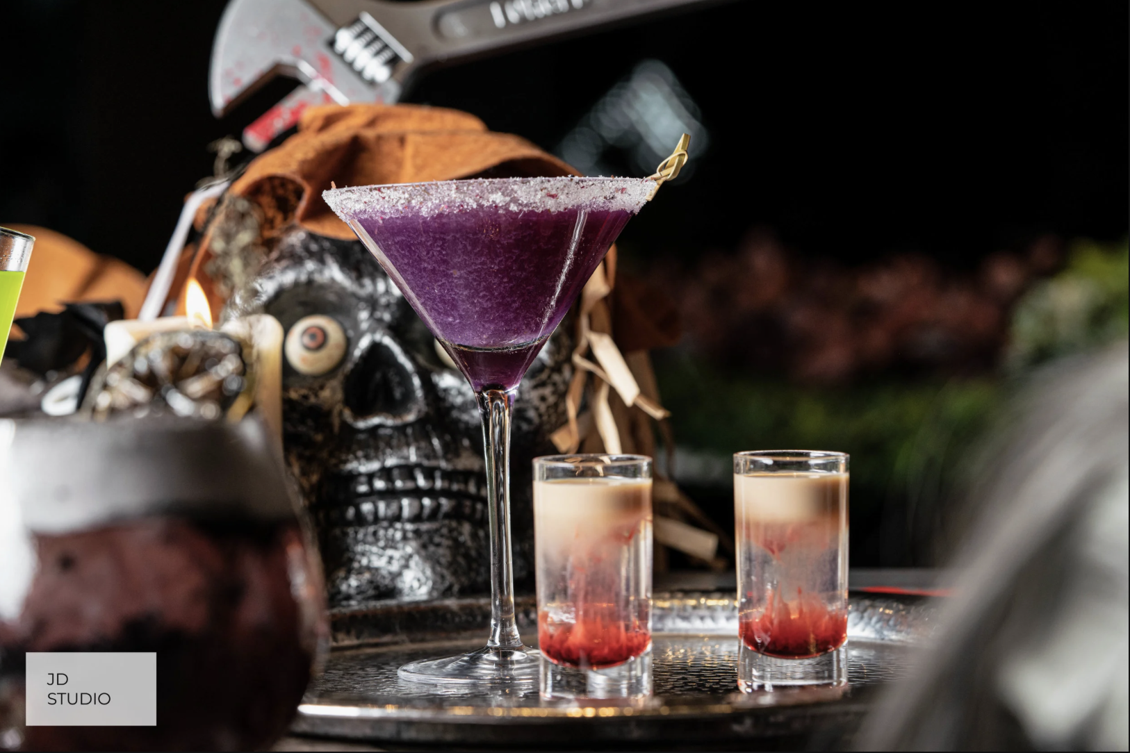 A purple cocktail in a martini glass with a sugar rim, accompanied by two shots with layered red and cream colors, on a decorative tray with Halloween themed decorations including a skull mask and a brown cloth in the background.