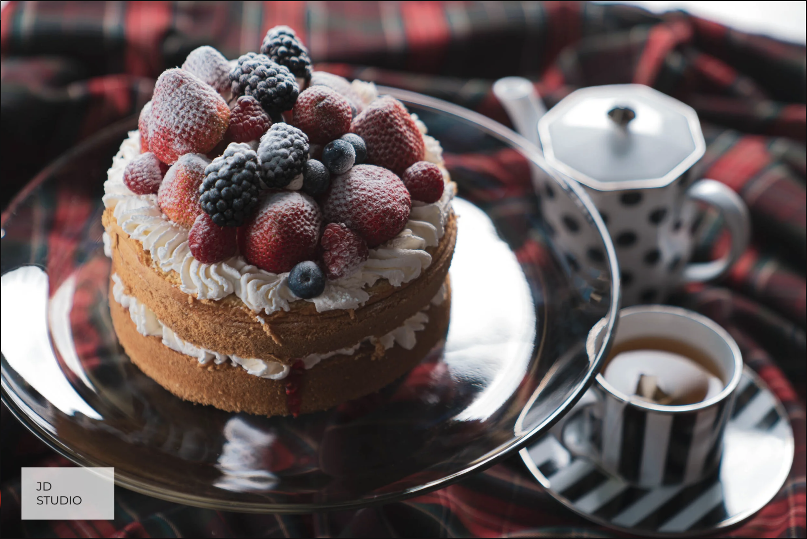 A layered cake topped with assorted berries, including strawberries, blackberries, blueberries, and raspberries, on a silver platter. In the background, there are a polka-dotted teapot and a striped cup of tea, set on a dark red plaid cloth.