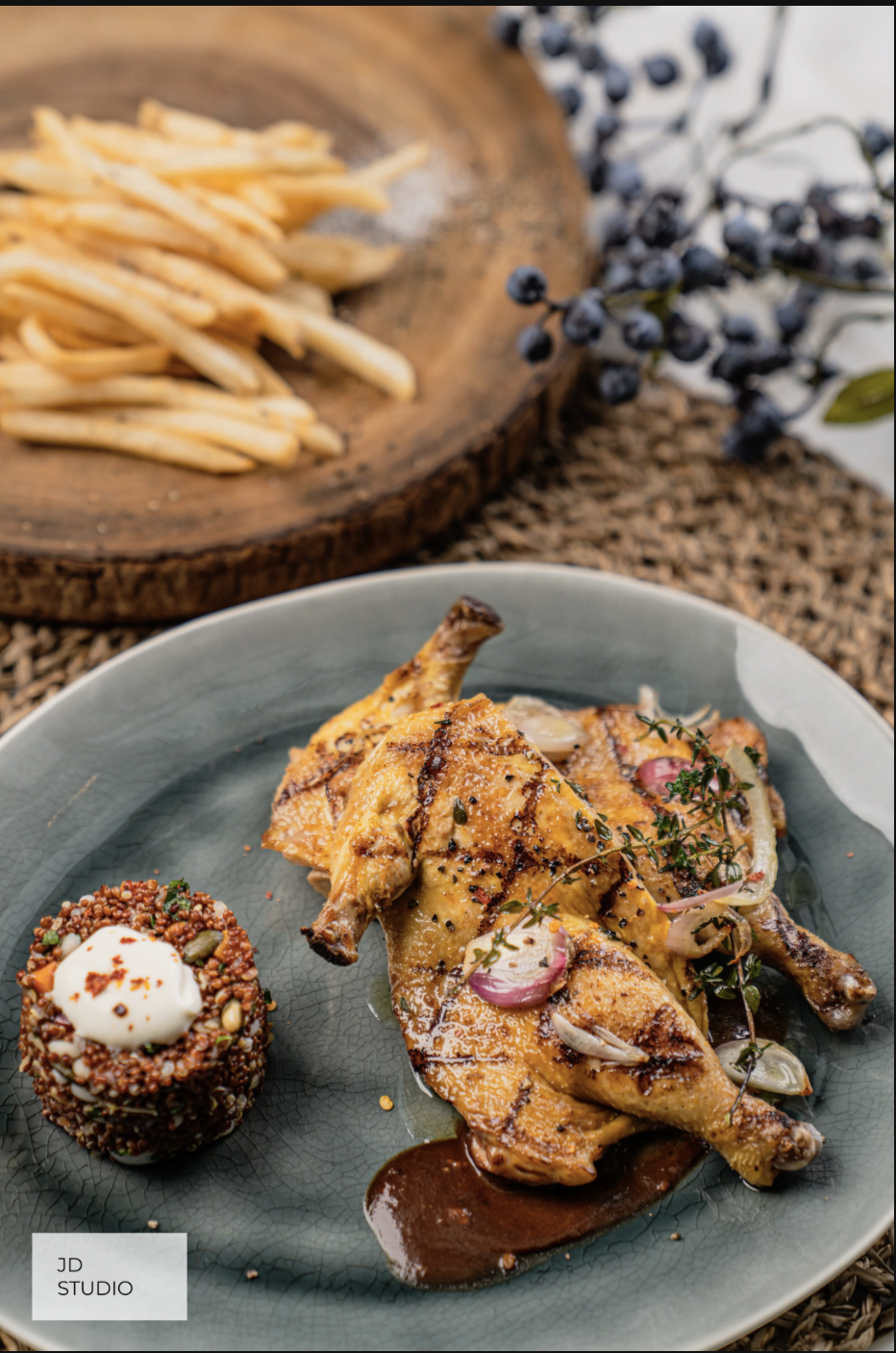 Grilled chicken served with a side of grain and herbs on a gray plate, with French fries on a wooden tray in the background, and black berries on a branch.