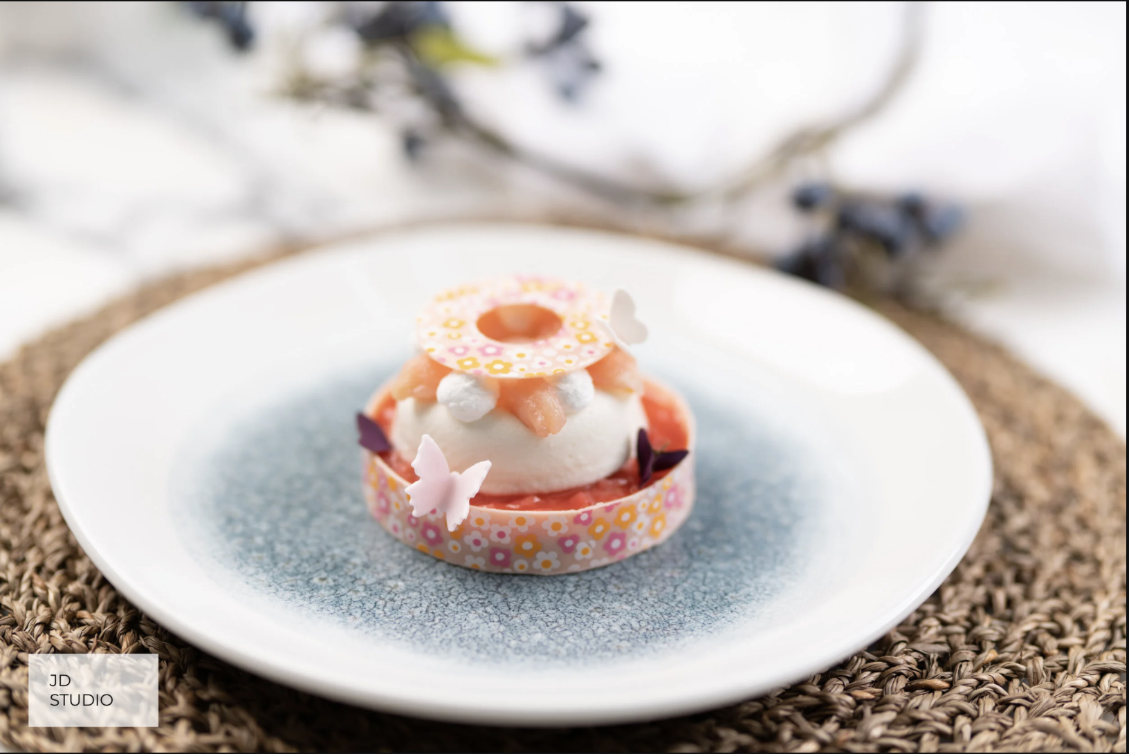 A small, ornate dessert with a pink and yellow floral patterned base, topped with a scoop of white ice cream or mousse, decorated with small white pearls, peach-colored fruit, butterfly-shaped decorations, and a decorative ring with a butterfly on to