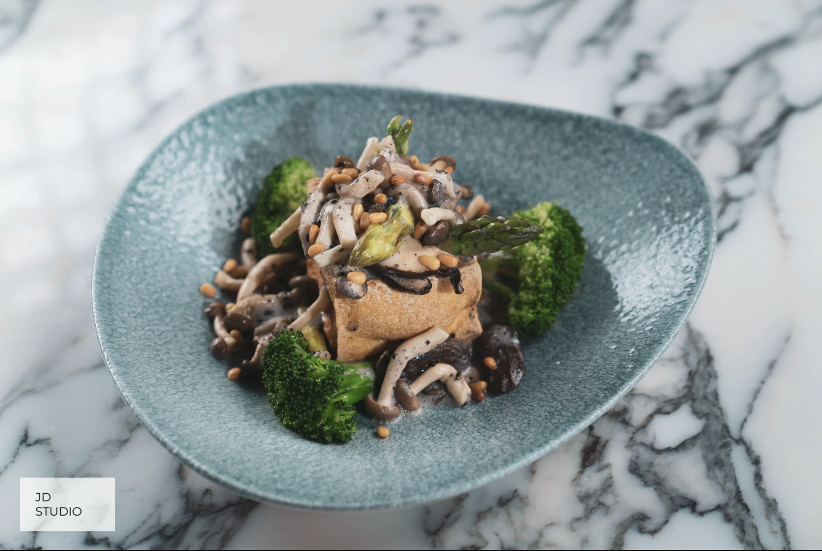 A vegetable dish with broccoli, mushrooms, and a stuffed pastry topped with mushrooms and pine nuts served on a blue textured plate on a marble surface.