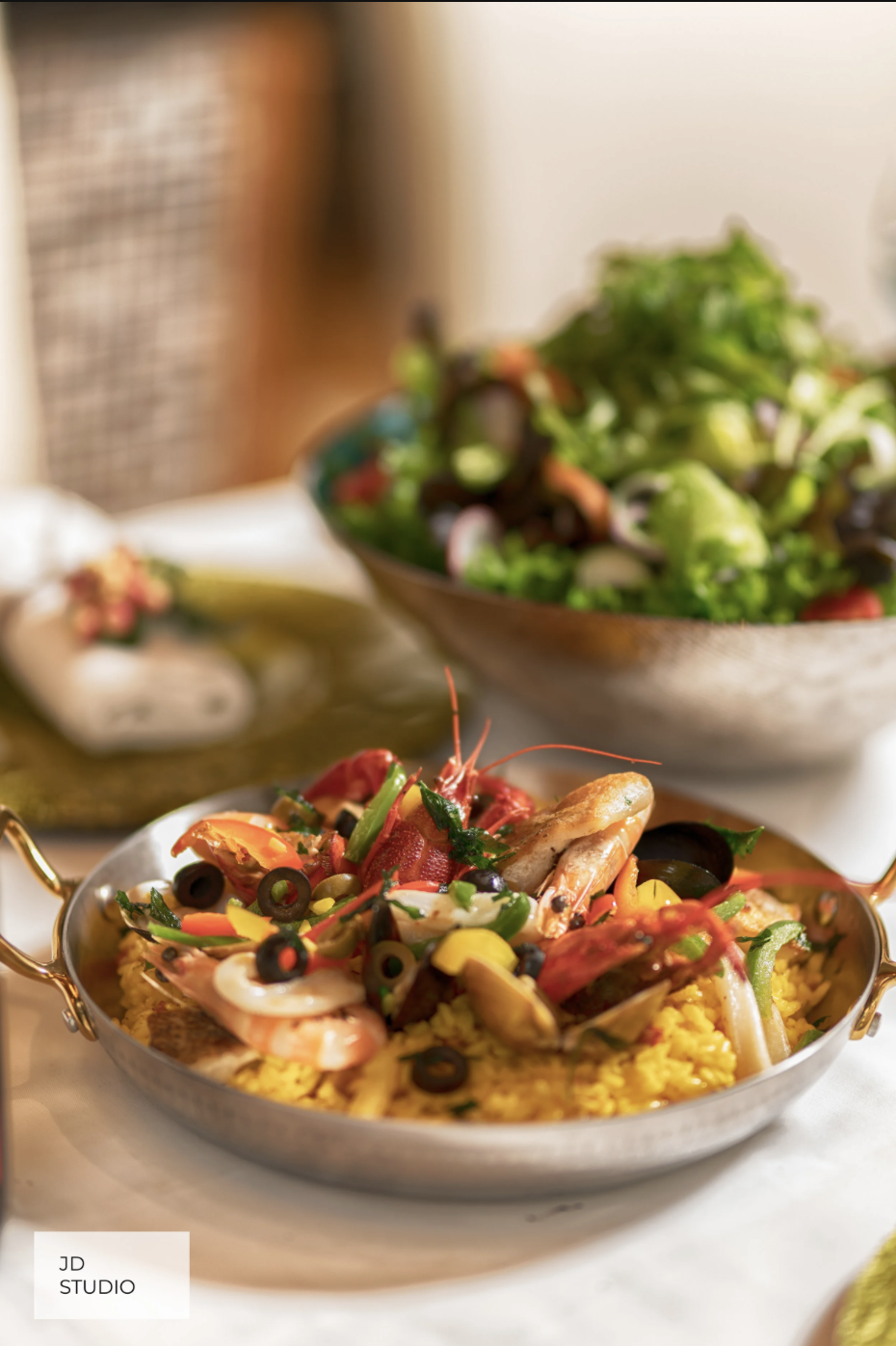 A seafood paella with shrimp, squid, and assorted vegetables served in a shallow metallic dish with a bowl of mixed salad in the background.