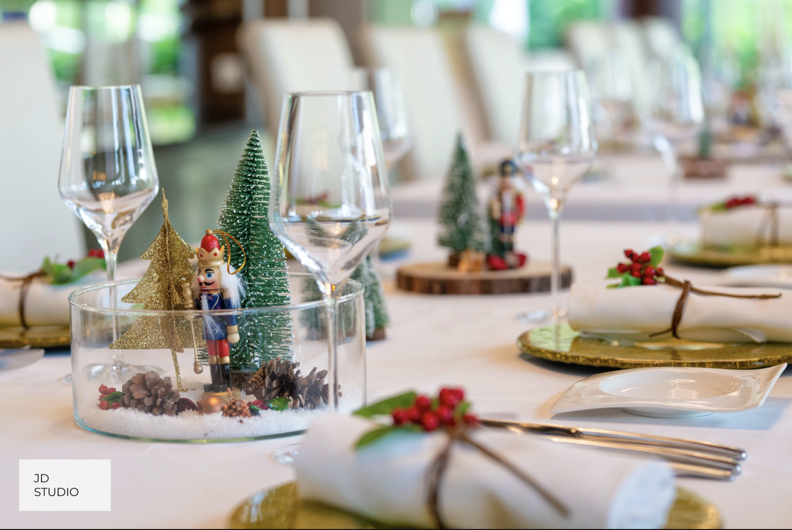 Decorated holiday table with Christmas centerpiece featuring miniature trees, nutcracker, pinecones, and snow, along with wine glasses, plates, napkins, and festive accents.