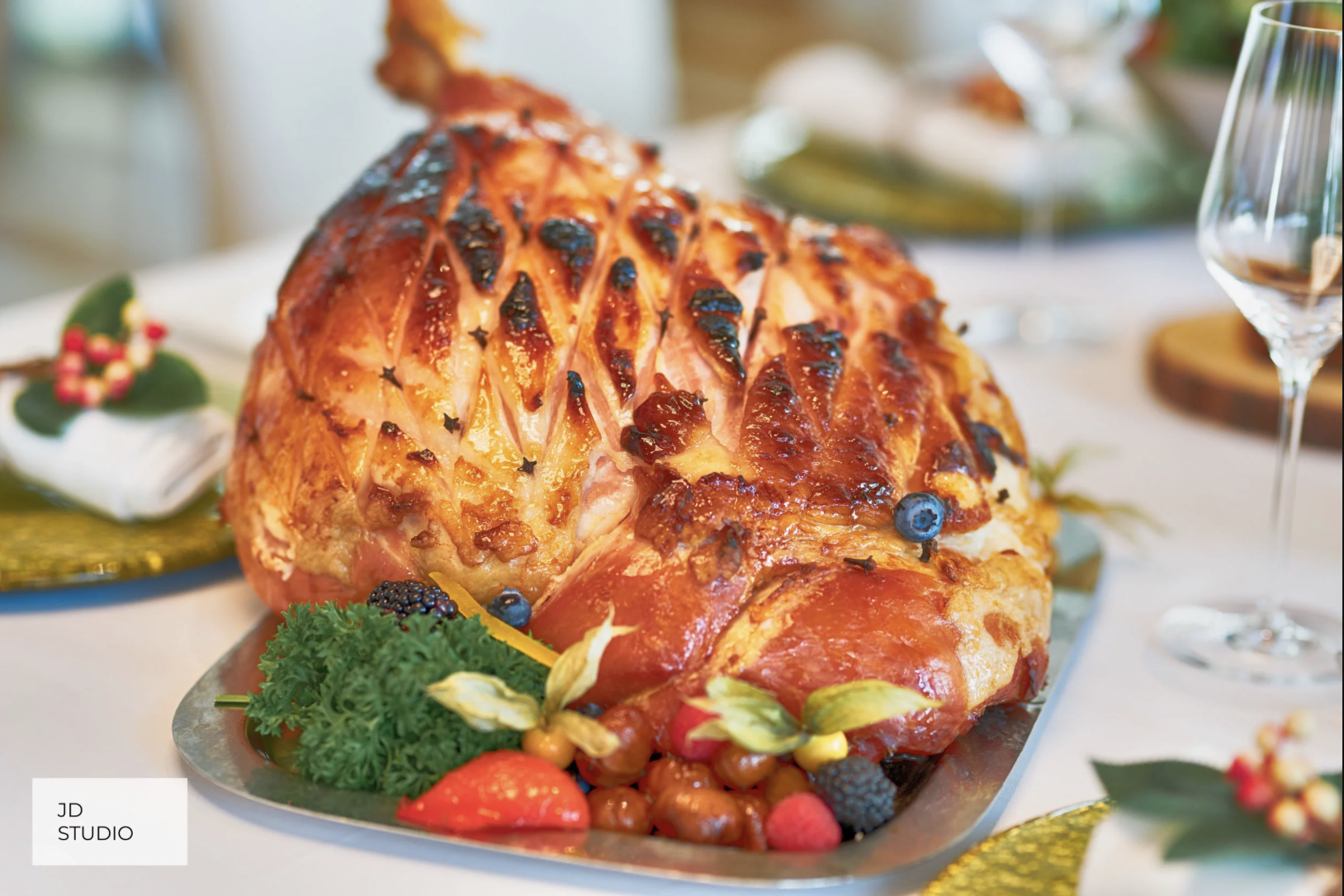 Decorated cooked ham with a honey glaze, garnished with blueberries, grapes, strawberries, blackberries, parsley, and other assorted fruits on a silver platter.