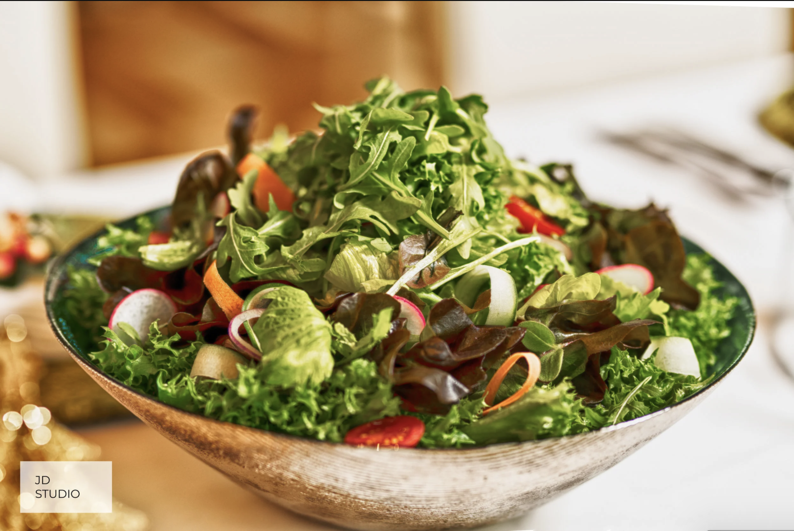 A large ceramic bowl filled with assorted fresh green salad greens, sliced radishes, cherry tomatoes, and shredded carrots on a white tablecloth.