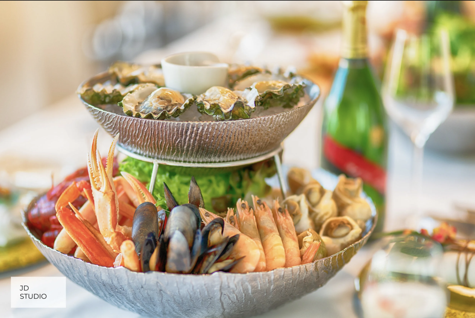 Tiered platter with oysters, shrimp, crab legs, and vegetables, served with a bottle of green beverage and wine glasses in the background at a seafood feast.