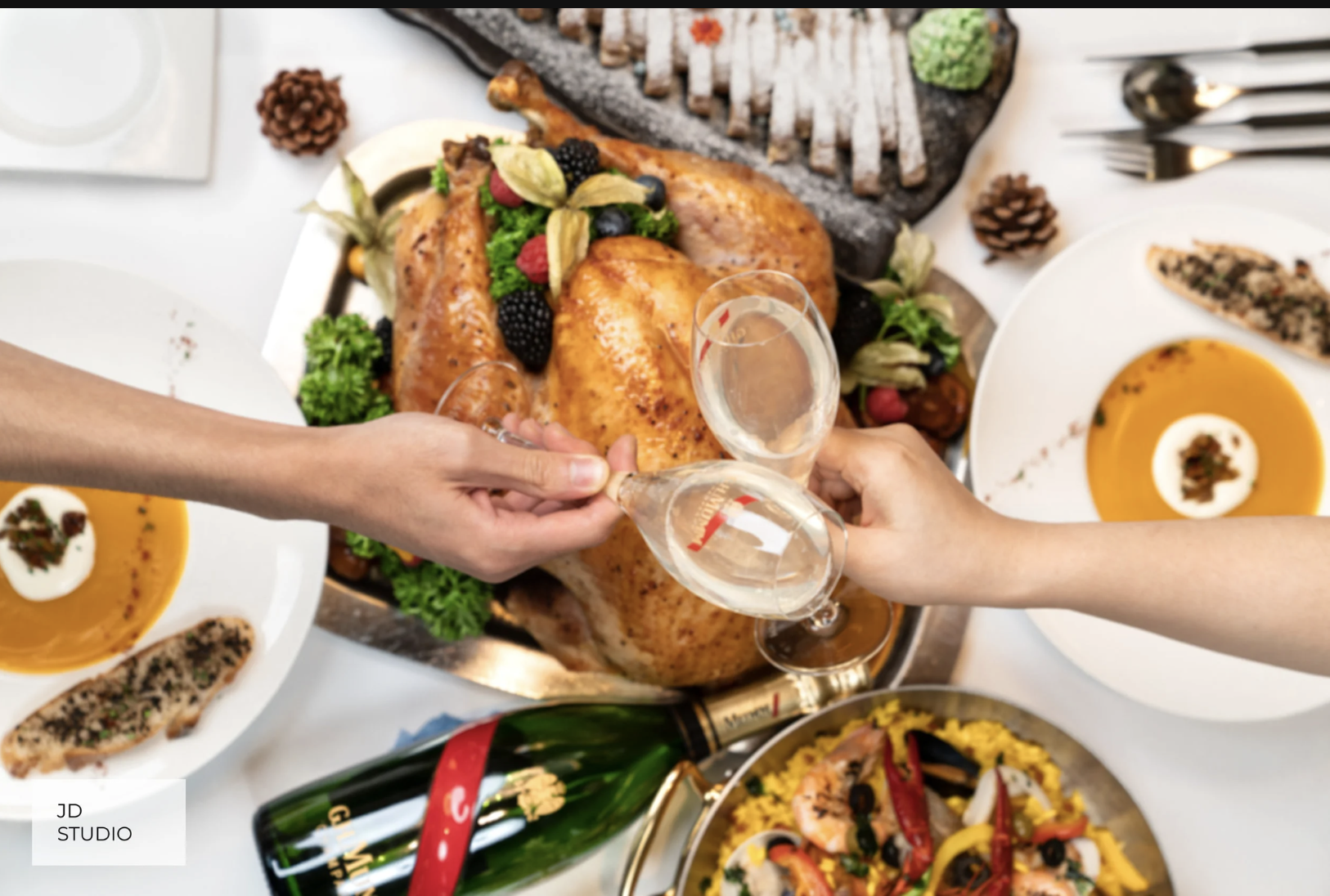 Two people clinking champagne glasses over a roast turkey with vegetables at a festive dinner table, surrounded by various dishes including soup, seafood, and side salads.