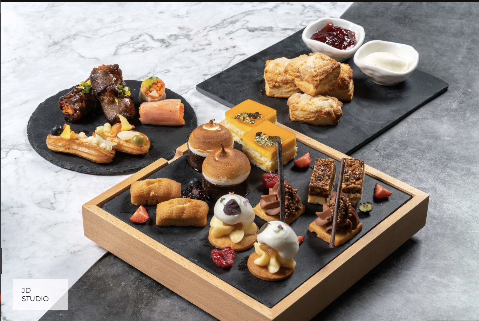 An assortment of desserts on a black tray, including mini tarts, layered cakes, and chocolate confections garnished with berries and edible gold leaf, with additional sweet treats on a black slate plate in the background.