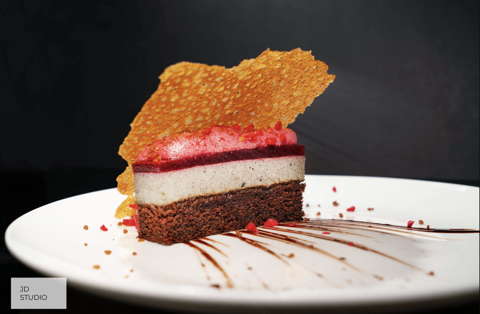 A slice of layered chocolate and berry mousse cake with a crispy caramel disc on top, served on a white plate with chocolate sauce drizzle and red crumbs, against a black background.