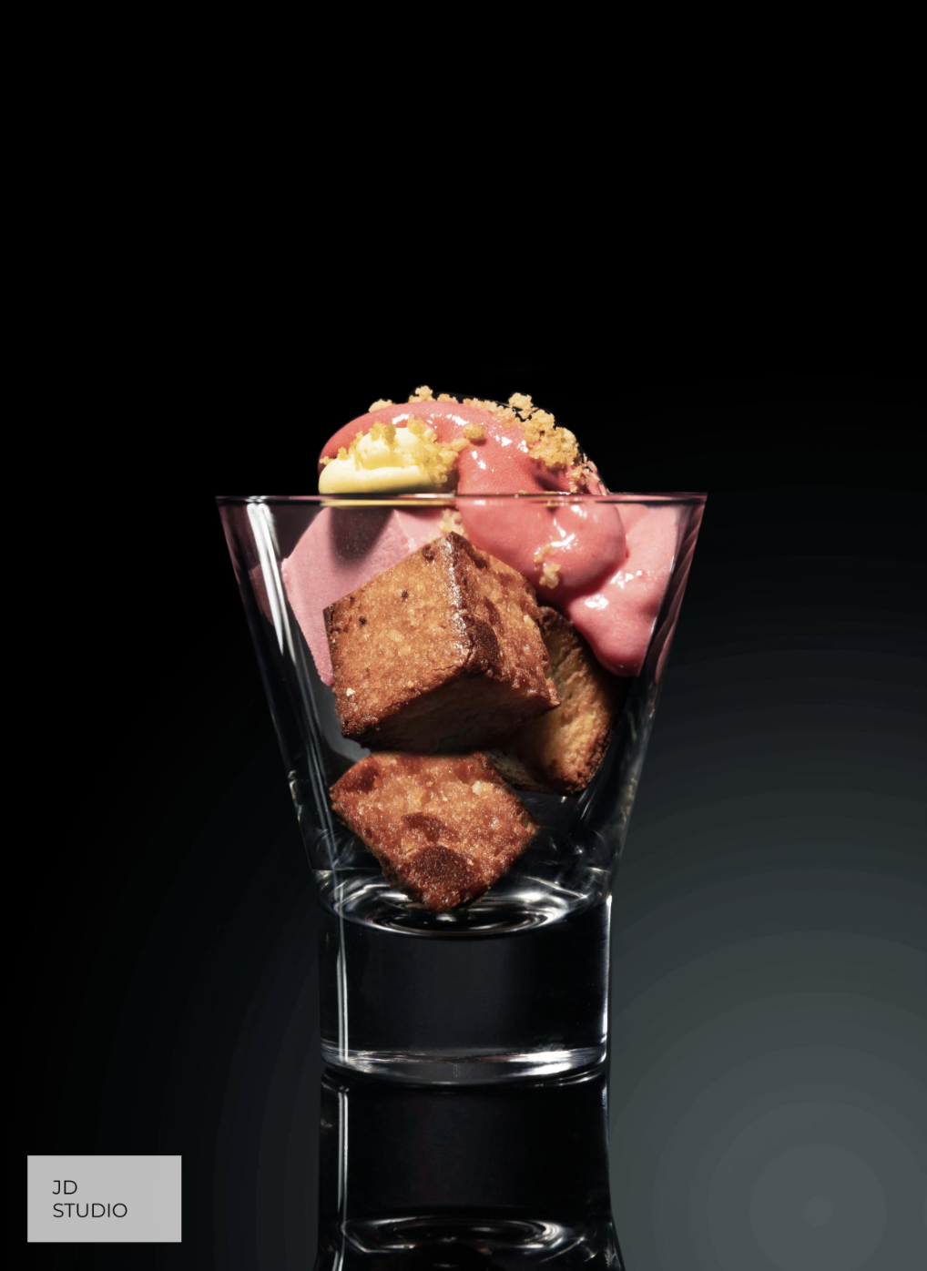 A clear glass cup with two pieces of cake and a scoop of pink and white ice cream on top, set against a black background.