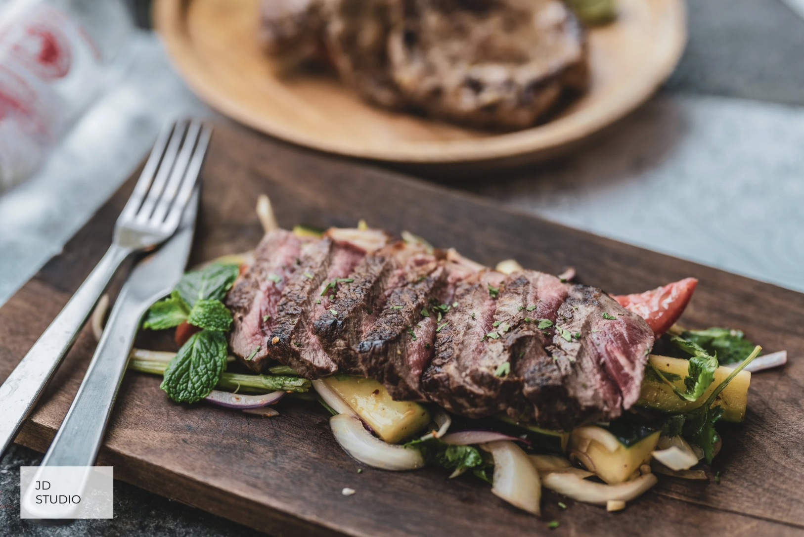 Sliced cooked steak on a bed of vegetables including onions, pineapple, and herbs, served on a wooden board.