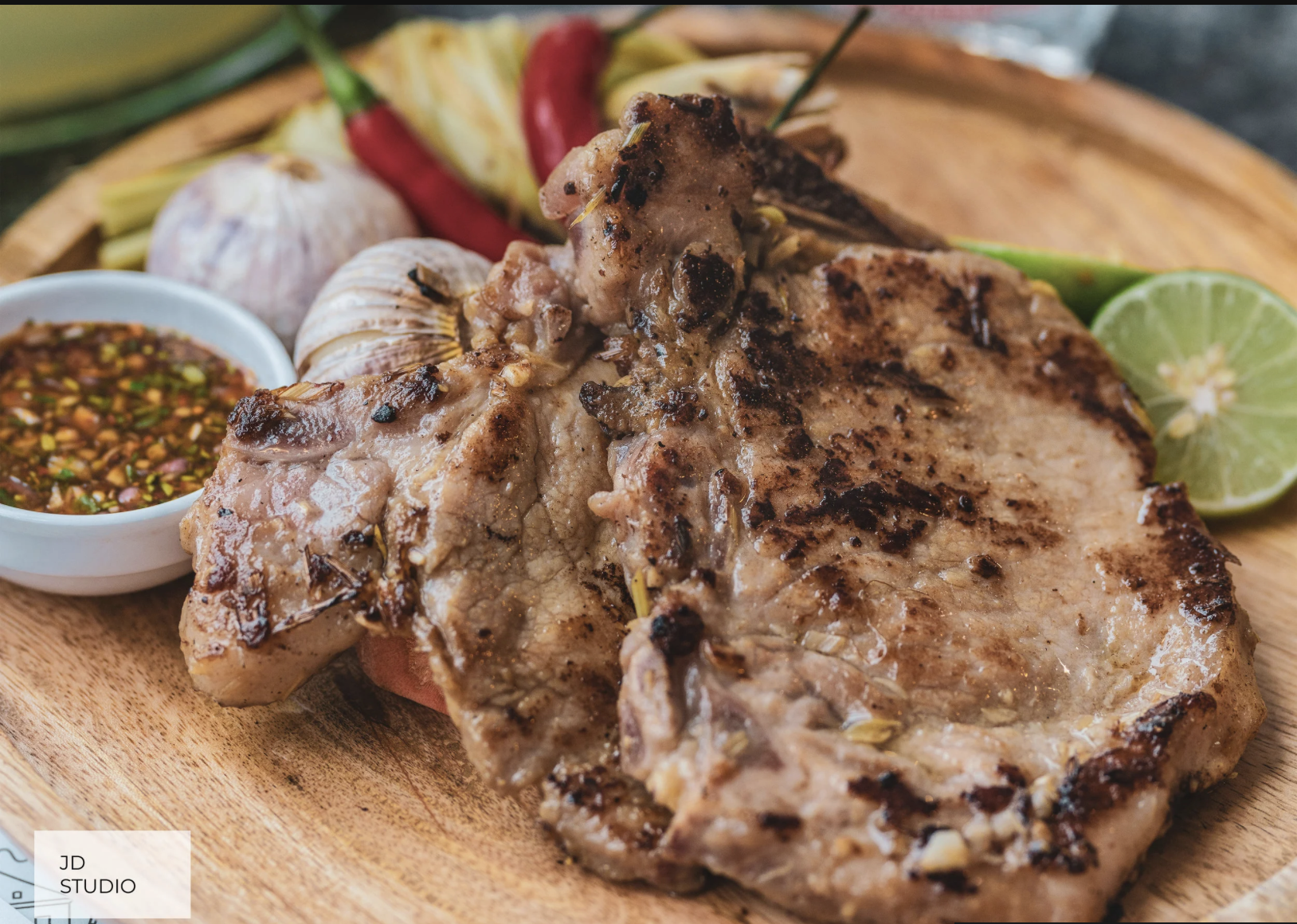 Grilled pork chop on a wooden plate with lime wedge, garlic, red chili peppers, and a small bowl of dipping sauce.