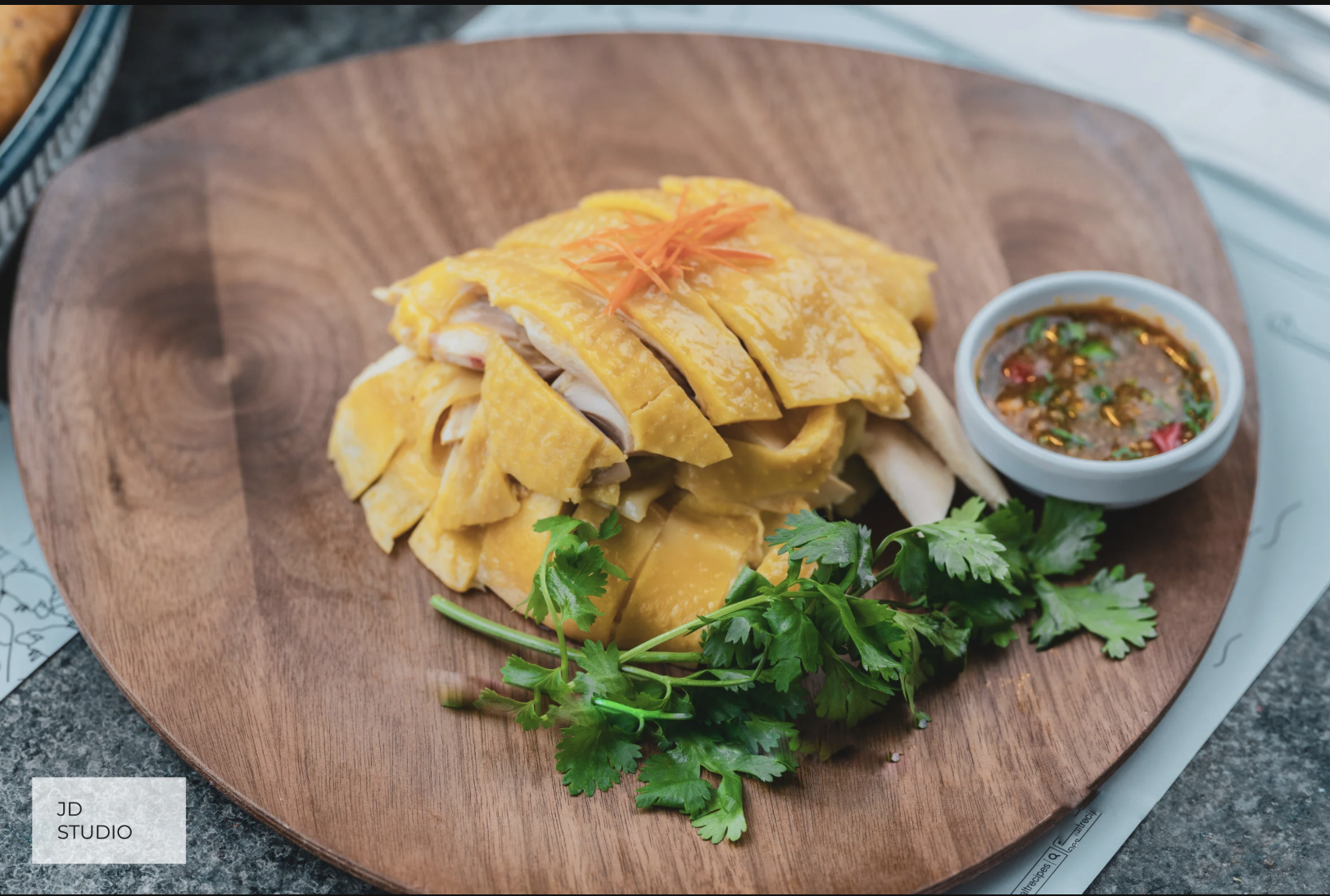Sliced roasted chicken served on a wooden platter garnished with fresh cilantro, alongside a small bowl of dipping sauce with chili and herbs.