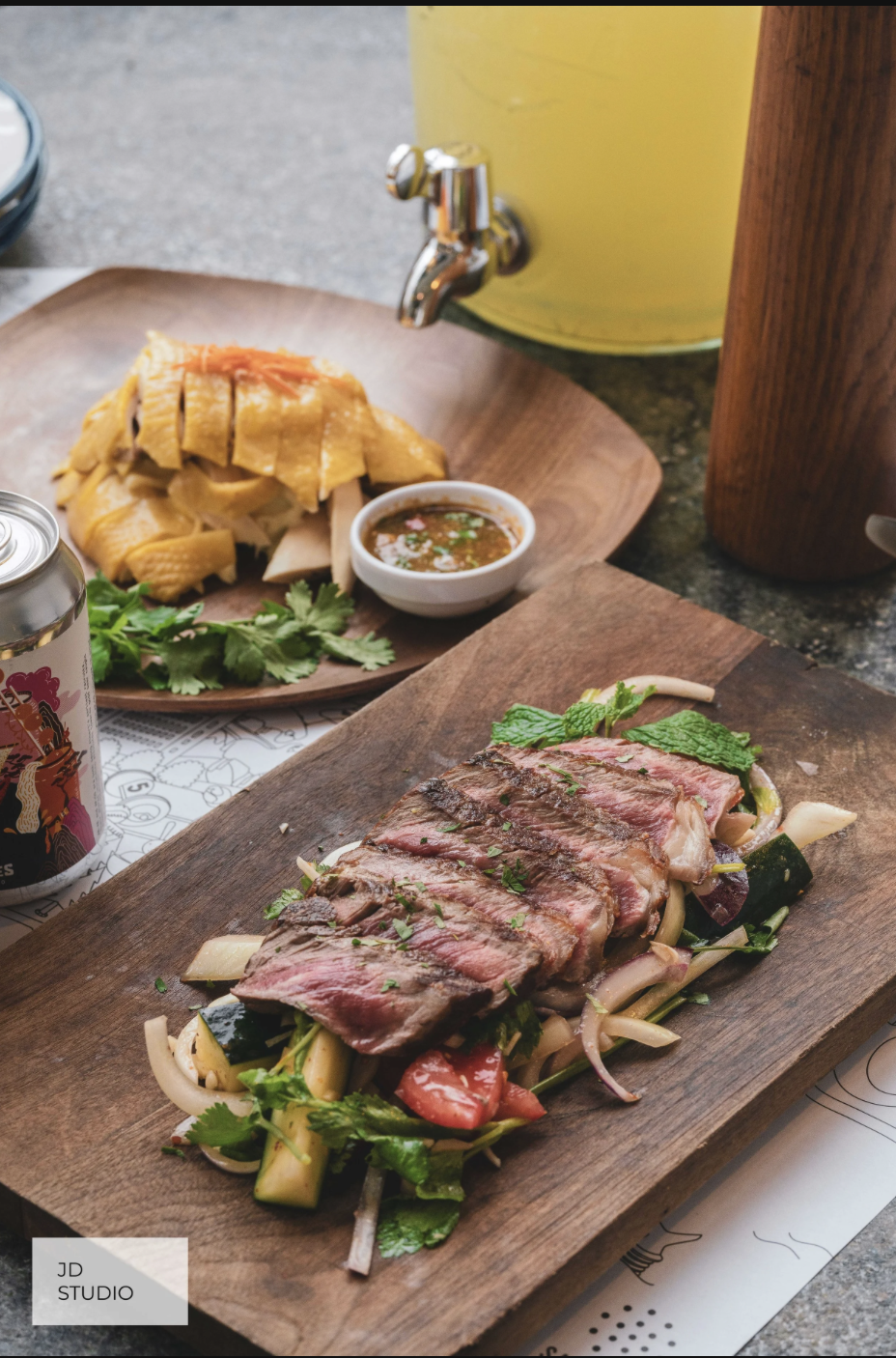 A wooden serving board with grilled steak, vegetables, and herbs. In the background, a wooden plate with sliced cooked chicken, cilantro, and dipping sauce, along with a drink can. Also visible is a yellow beverage dispenser and a paper placemat with