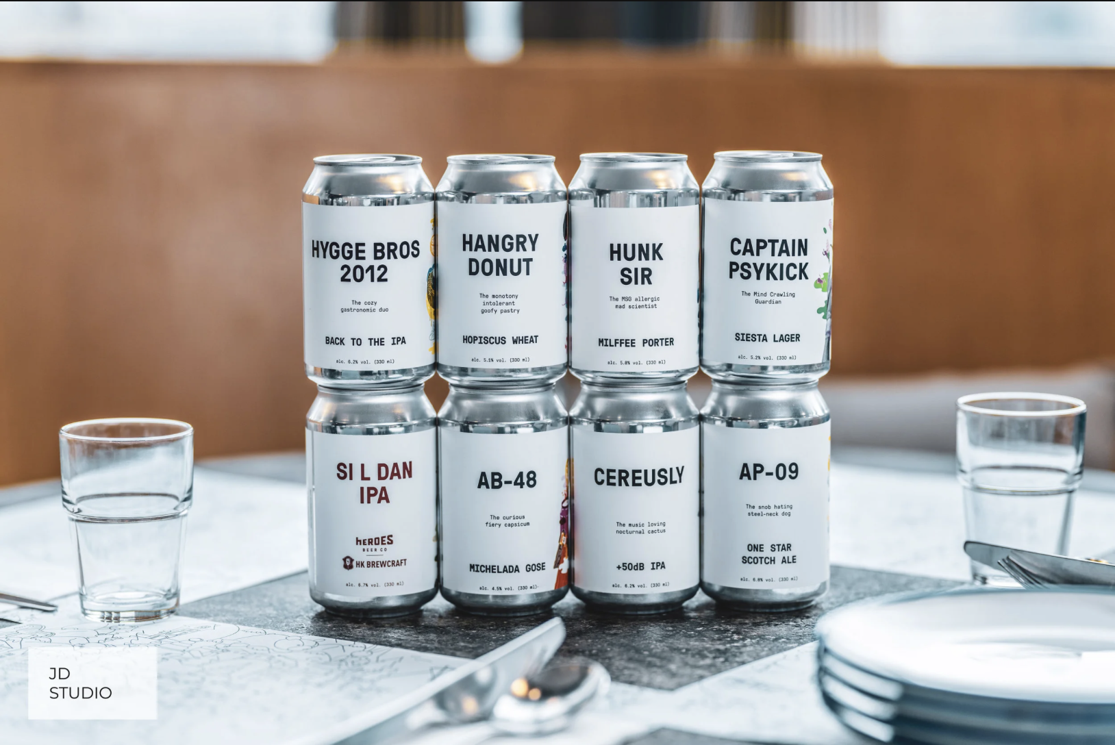 Stack of eight beer cans with creative labels on a dining table, with glasses and plates nearby.