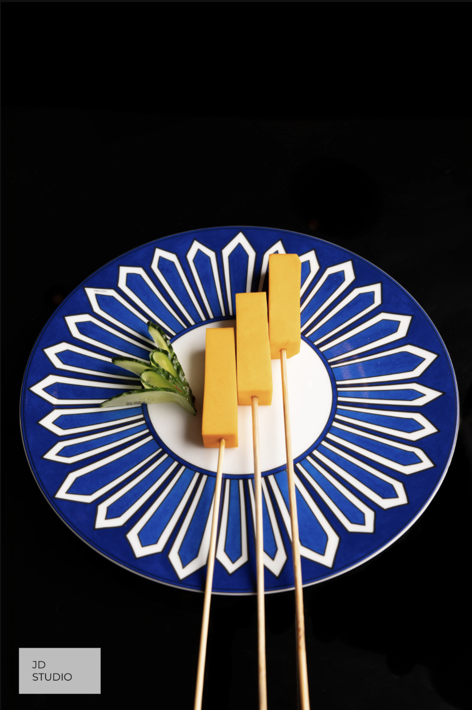 Slices of yellow cheese on wooden skewers placed on a decorative blue and white plate with cucumber garnish.