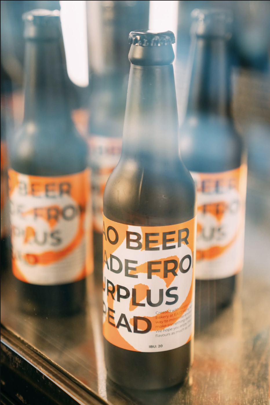 Multiple dark beer bottles with orange and white labels that say 'NO BEER MADE FROTHER PLUS DEAD' on a reflective surface.
