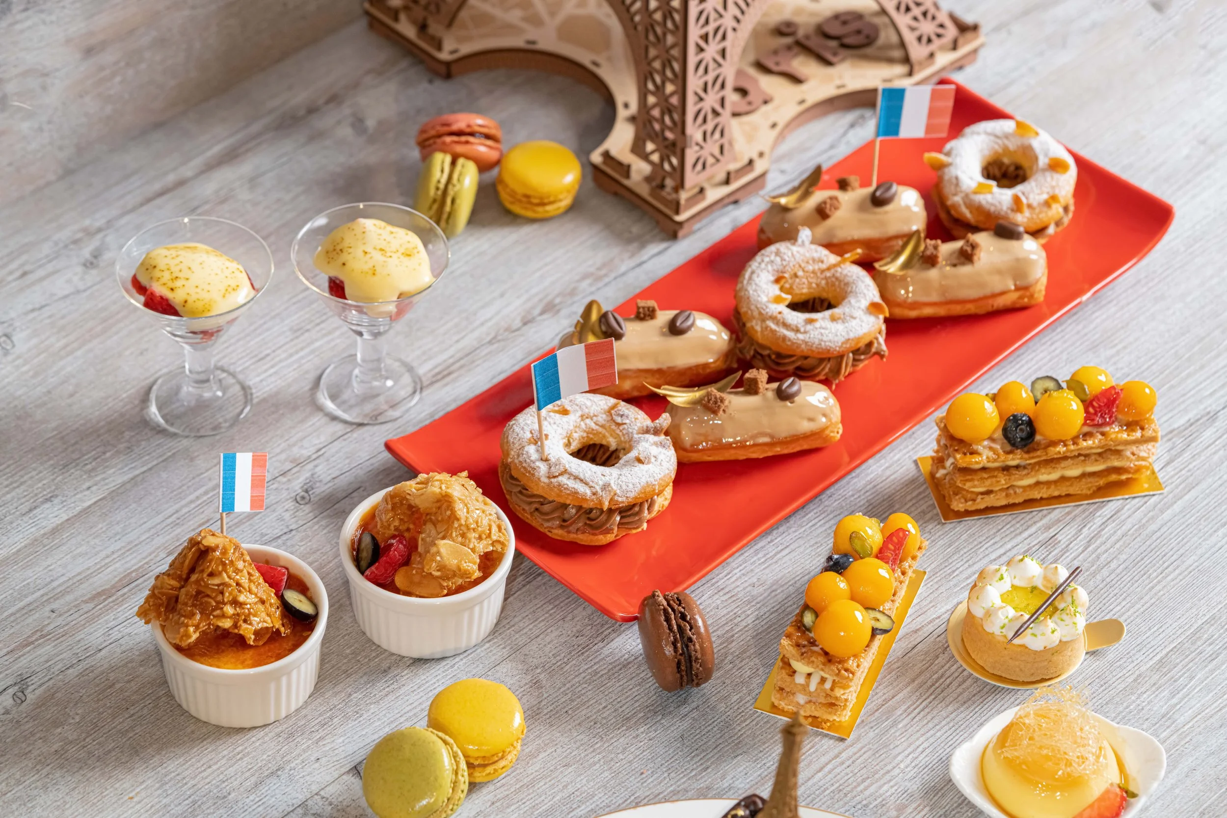 A table with various French desserts and macarons, including éclairs, a layered pastry with yellow toppings, fruit-topped petite tarts, and two small glasses with yellow custard desserts. There are also macarons in green and yellow, small flags of Fr
