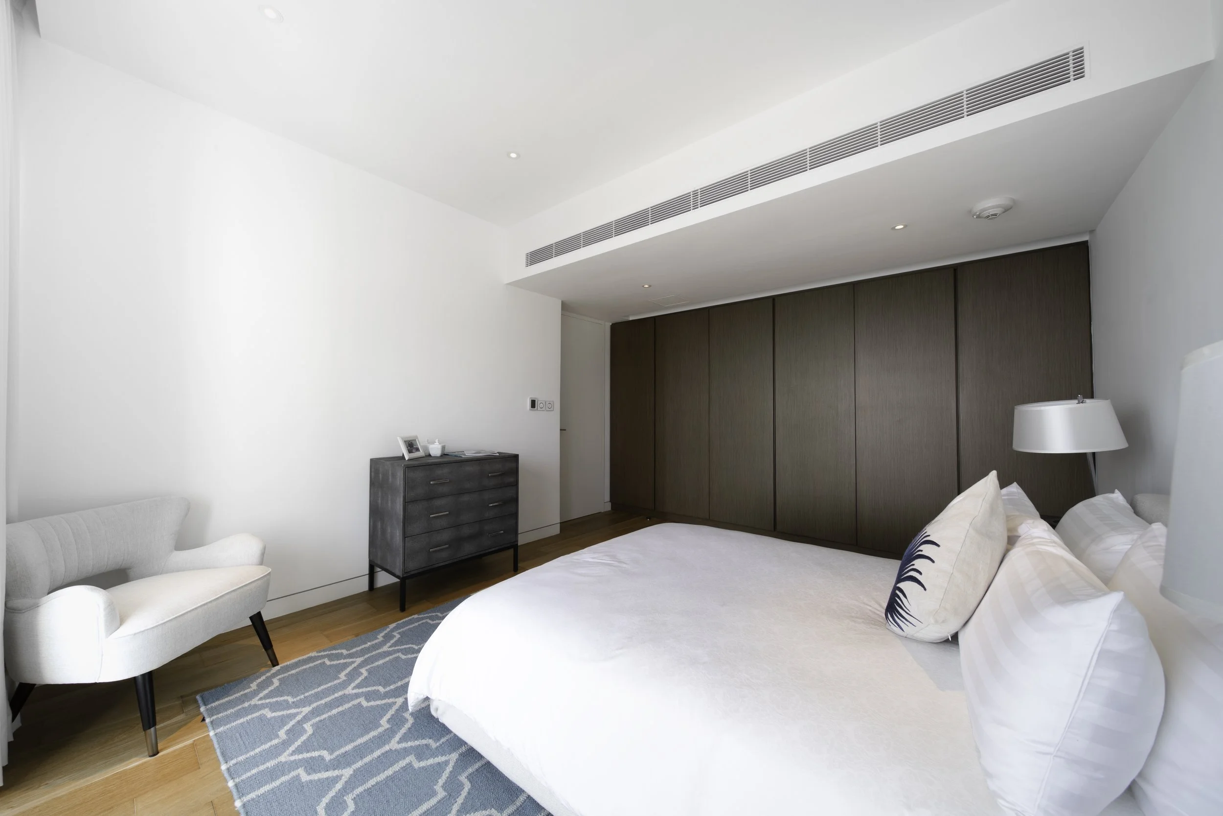 Modern bedroom with white bed, decorative pillows, bedside lamp, gray armchair, and dark wooden closet doors.