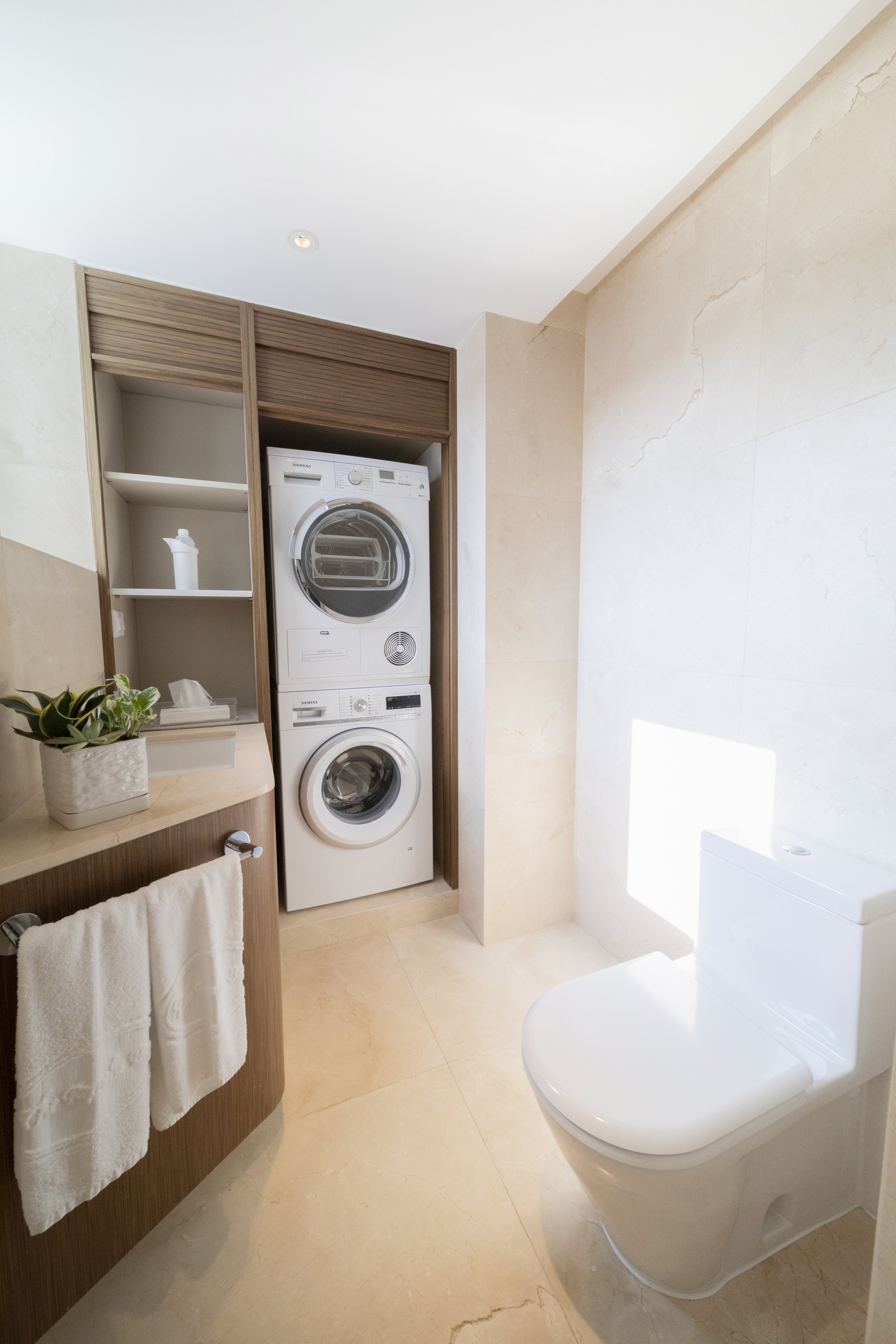 A modern bathroom with a stacked washer and dryer, a white toilet, and a sink with a towel rack and decorative plant.