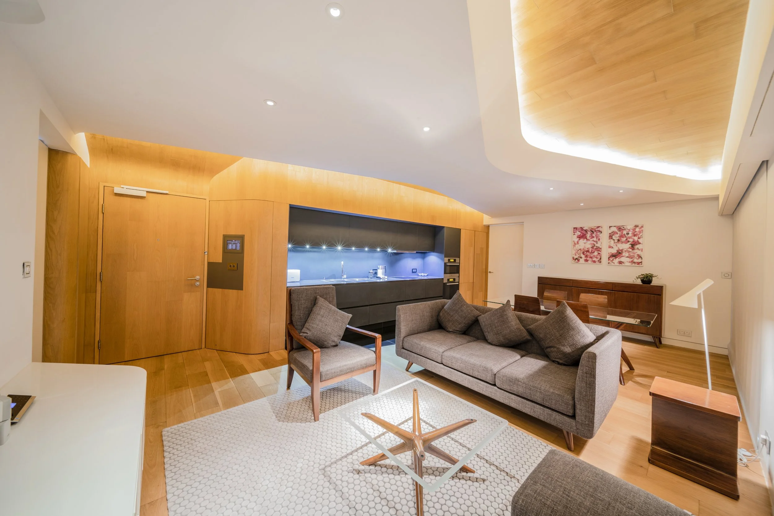 Modern living room with beige sofa and wooden armchair, glass coffee table, wooden side table with lamp, and a wooden cabinet with two pink floral paintings above. Kitchen area with dark cabinetry and backsplash in the background, ceiling with cut-ou