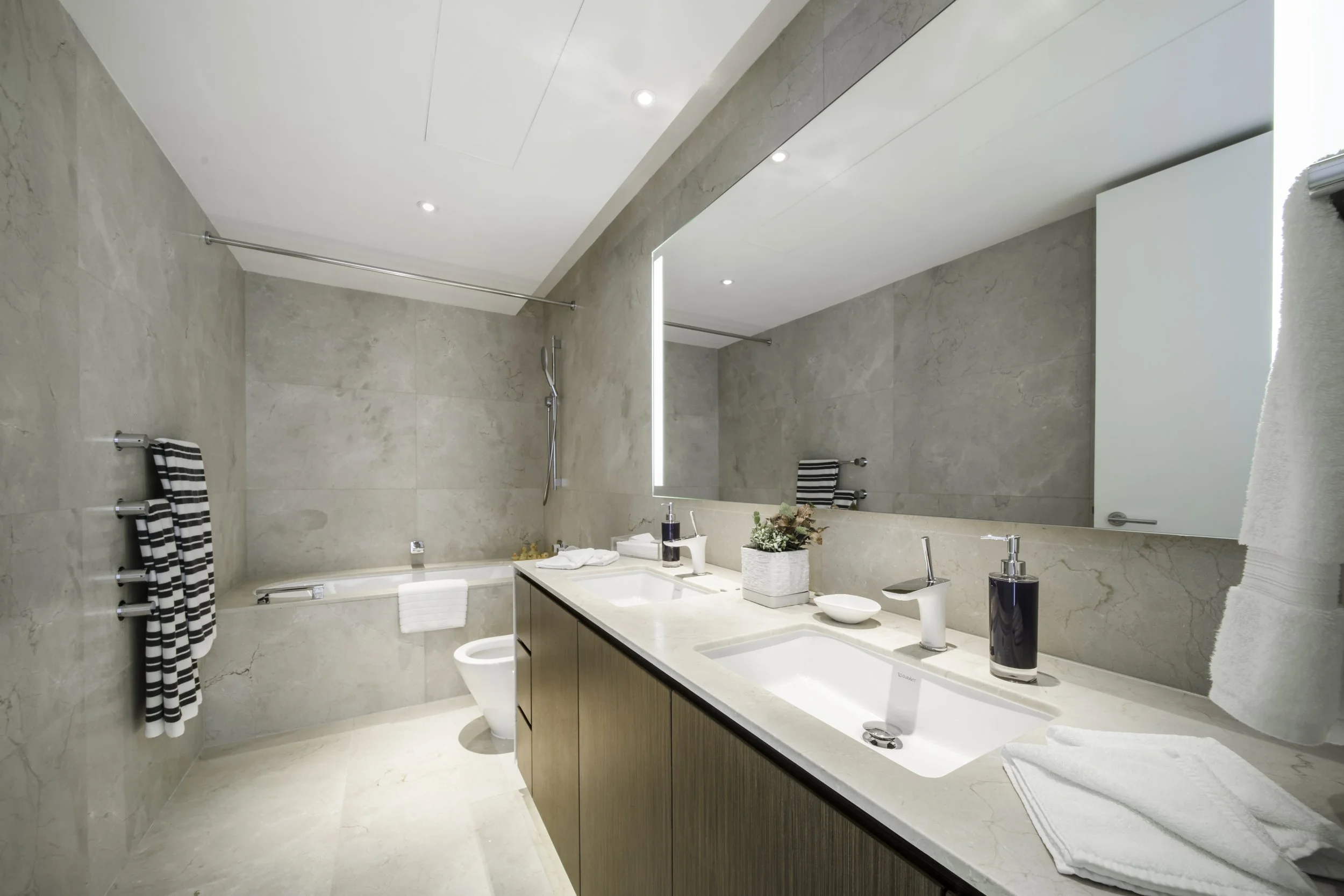 Modern bathroom with dual sinks, large mirror, towel racks, bathtub, and neutral-colored tile walls and floor.