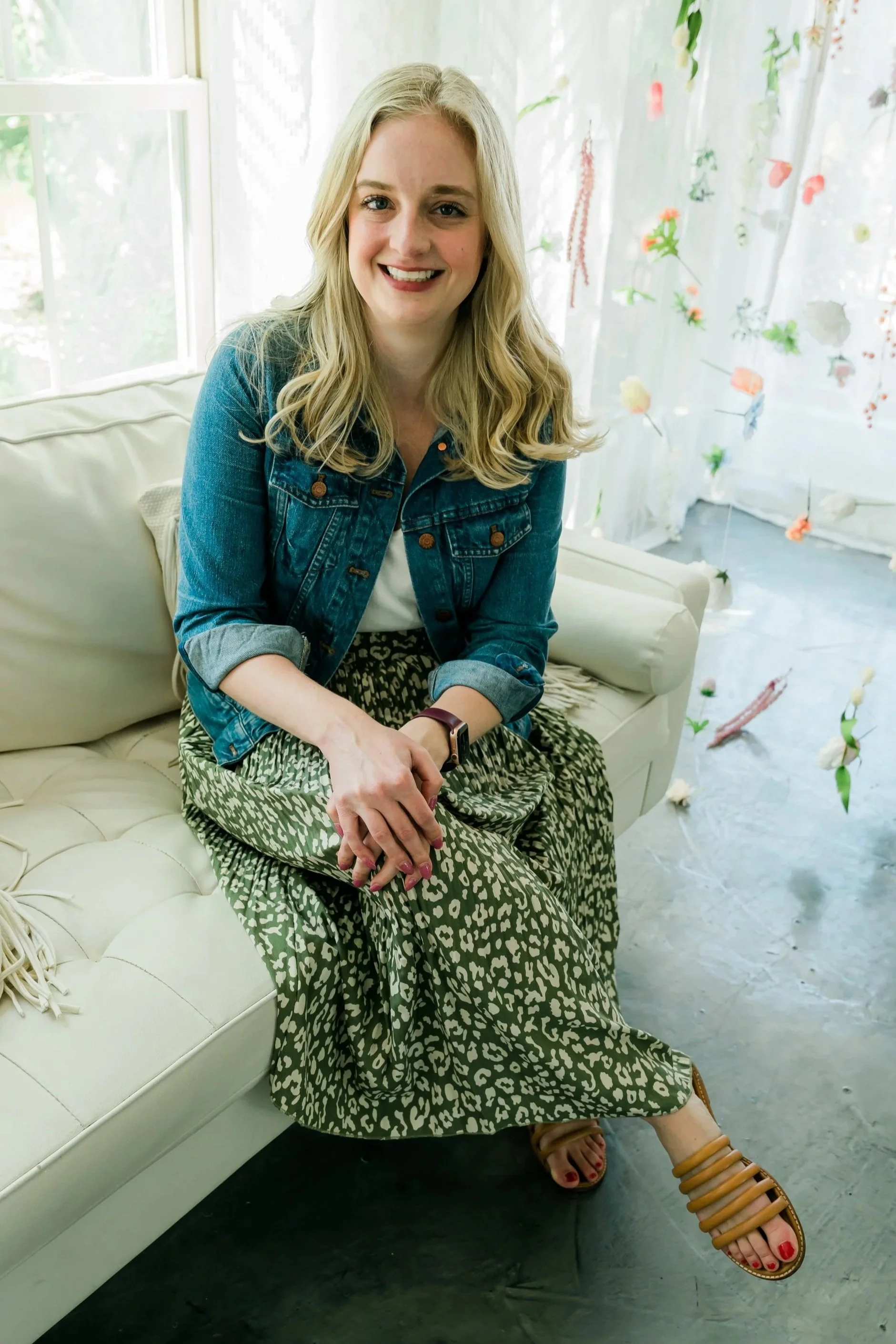 A woman with long blonde hair smiling, wearing a denim jacket and patterned pants, sitting on a white couch in a bright room with floral curtains.