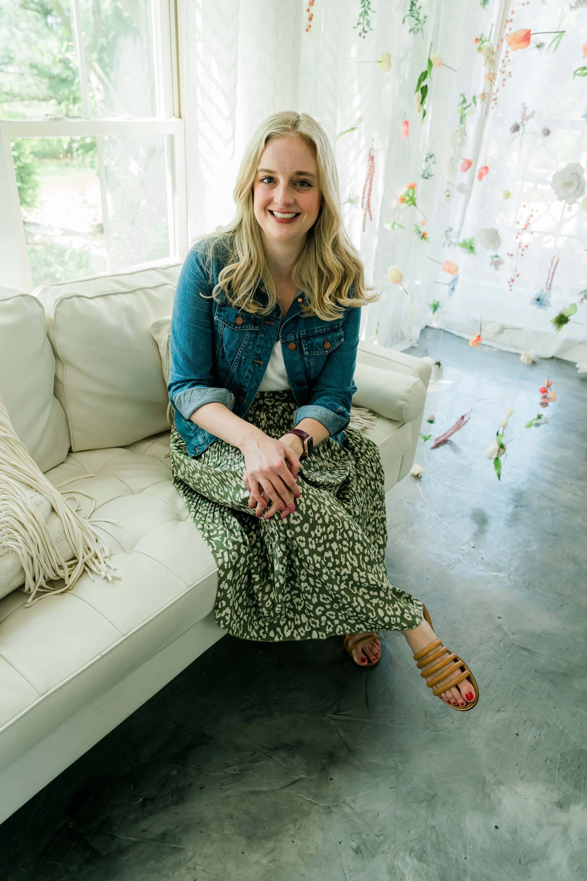 A woman with blonde hair smiling, sitting on a cream-colored sofa in a bright room with floral curtains, green foliage outside the window, and a concrete floor.