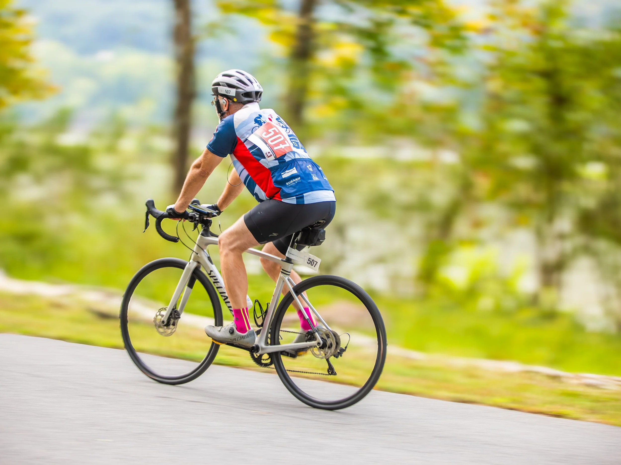 Person riding a mountain bike on a paved path in a park with green trees and grass, wearing a white helmet and colorful cycling uniform.