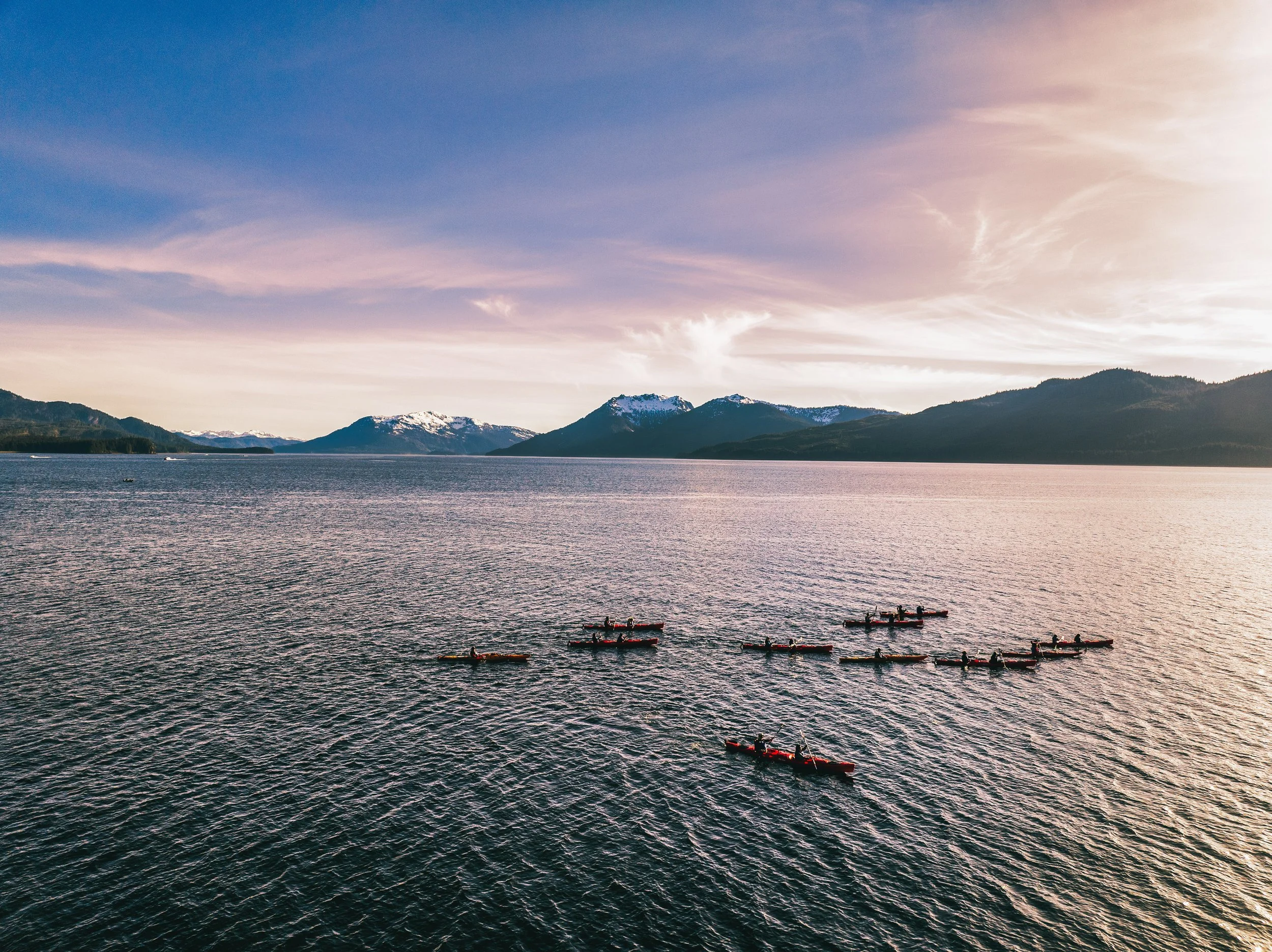 A scenic view of a large body of water with several kayakers paddling in a line, mountains with snow-capped peaks in the background, under a sky with streaks of pink and blue clouds.