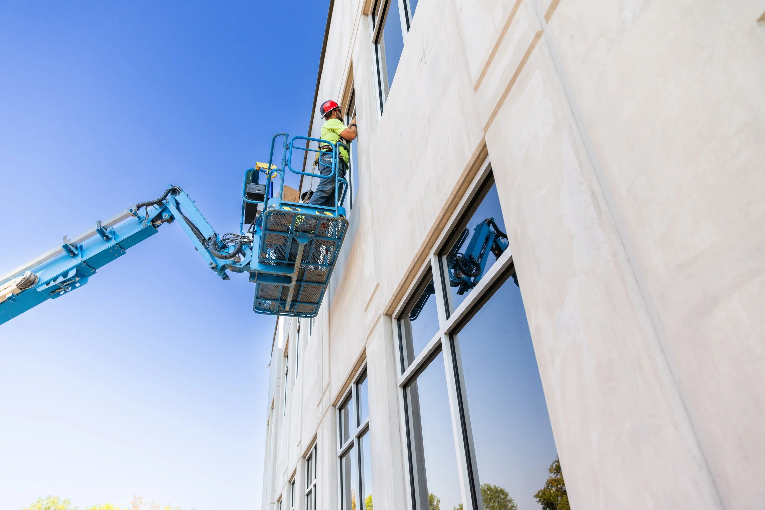 A construction worker wearing a red helmet and neon green shirt is standing on a blue hydraulic lift, working on the exterior of a modern building with large windows and a light-colored facade against a clear blue sky.