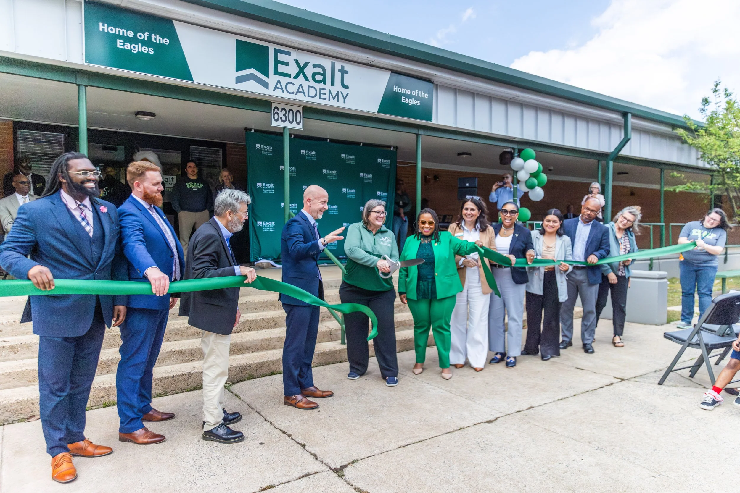 A group of diverse people attending a ribbon-cutting ceremony in front of an educational center called Exalt Academy, with a banner reading 'Home of the Eagles' and green, white, and black balloons.