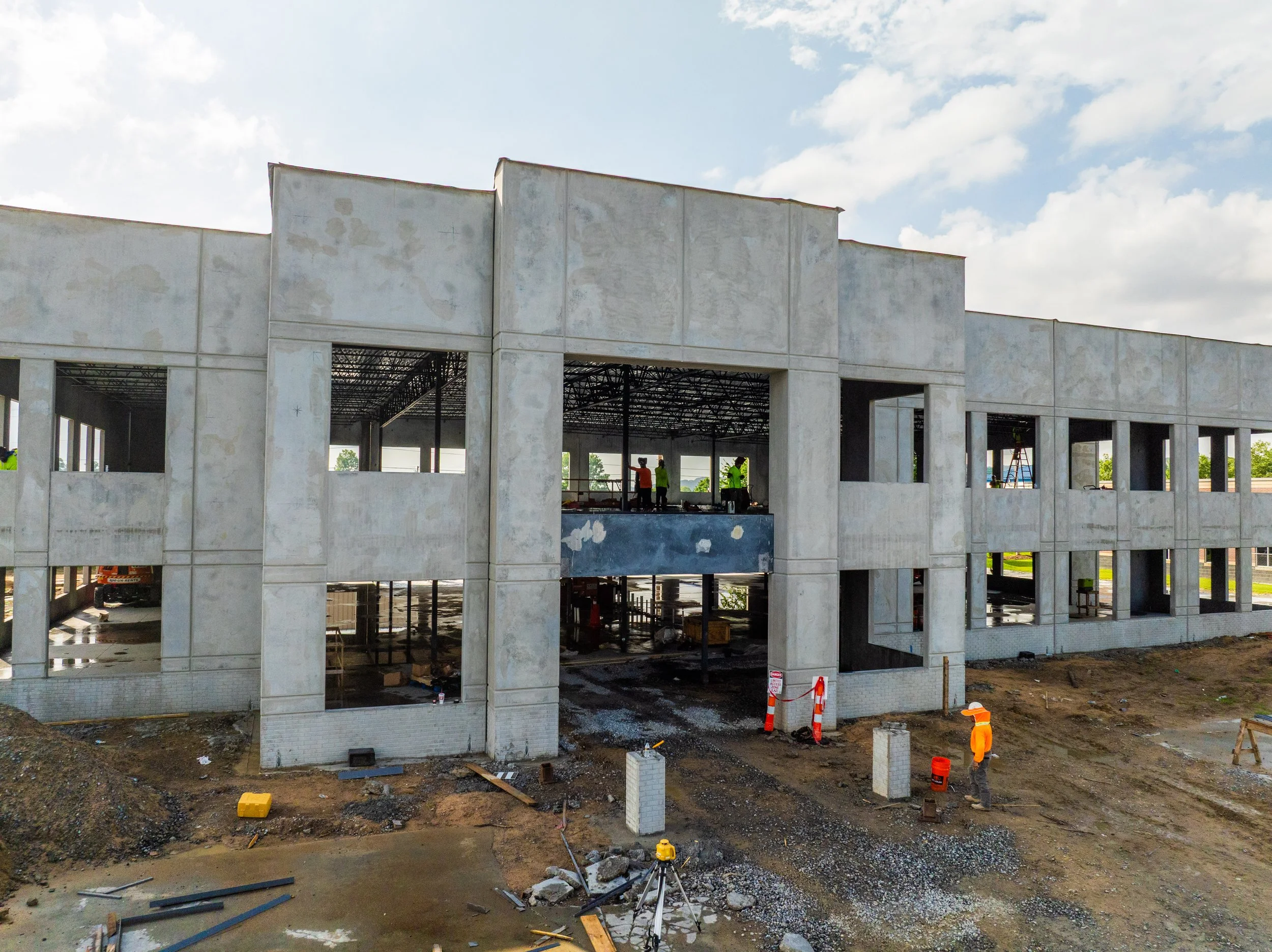 Construction workers building a multi-story concrete structure with open walls and large window openings, surrounded by construction equipment and dirt ground.