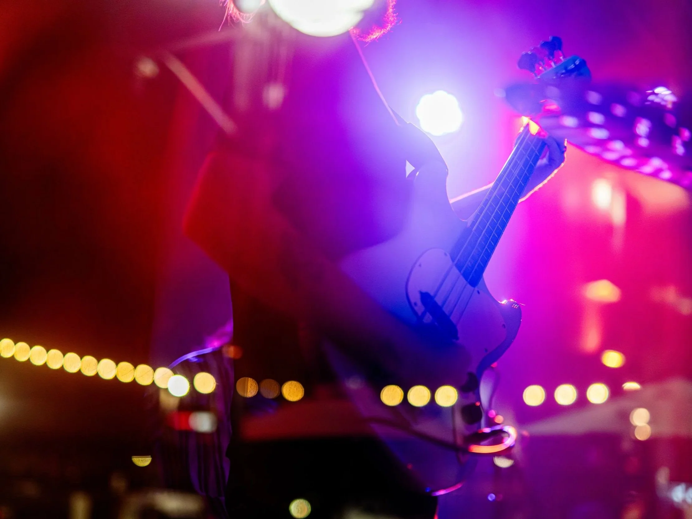 Person playing electric guitar on stage with colorful purple and pink lights.
