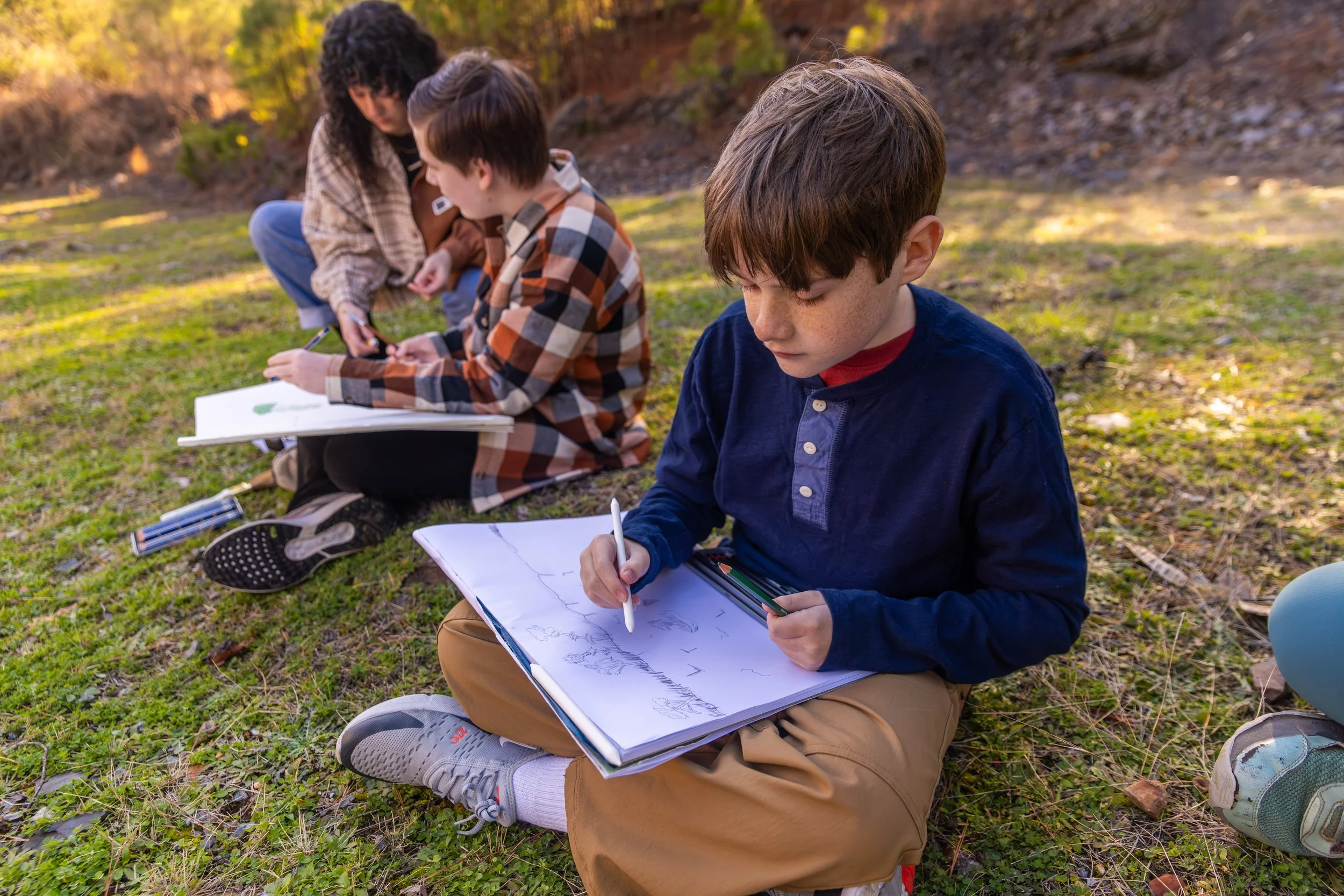 Two young boys and an adult sitting on grass outdoors, drawing and writing in sketchbooks, with trees and autumn foliage in the background.