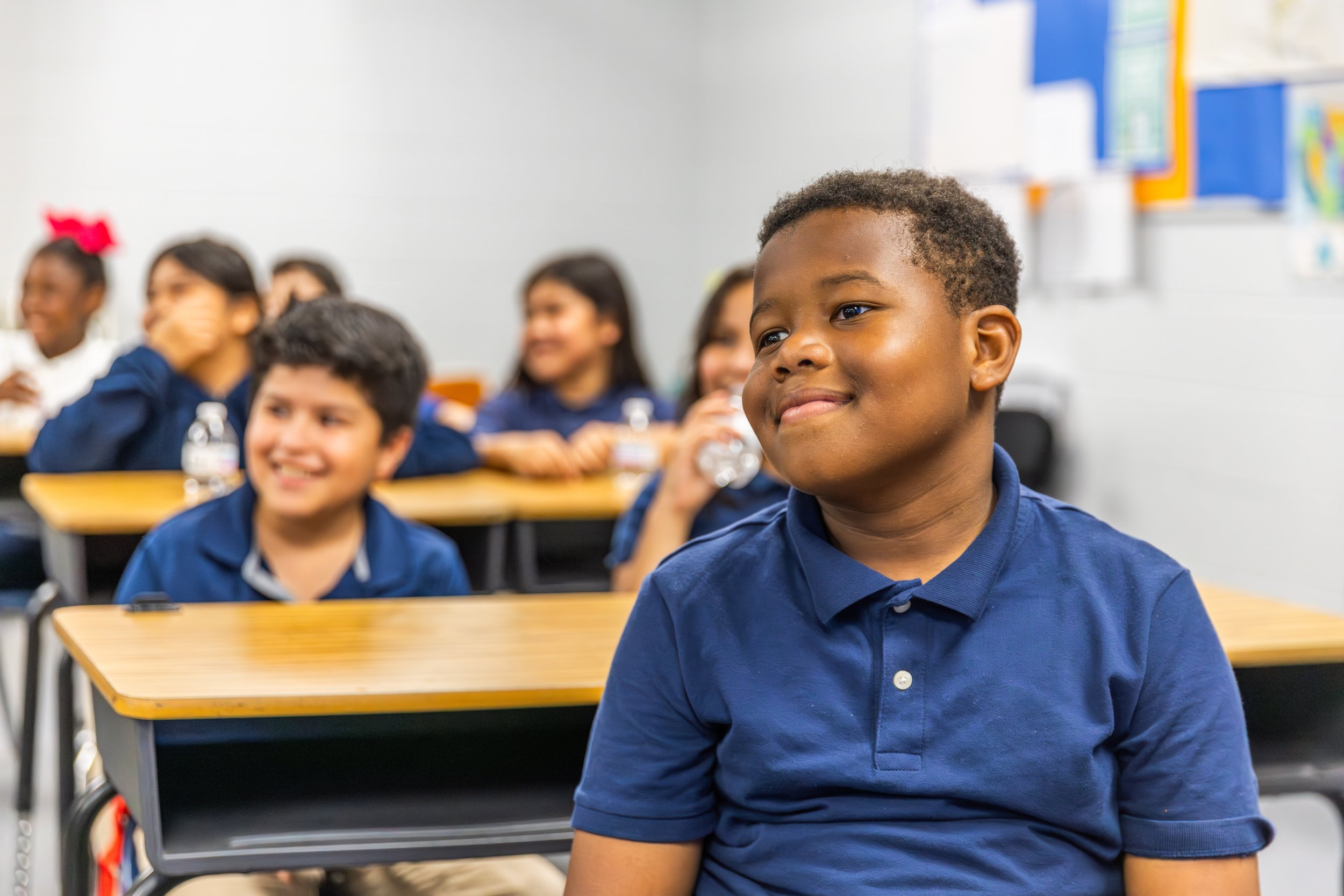 Children sitting at desks in a classroom, with a cheerful boy in a blue polo shirt in the foreground and other children smiling in the background.