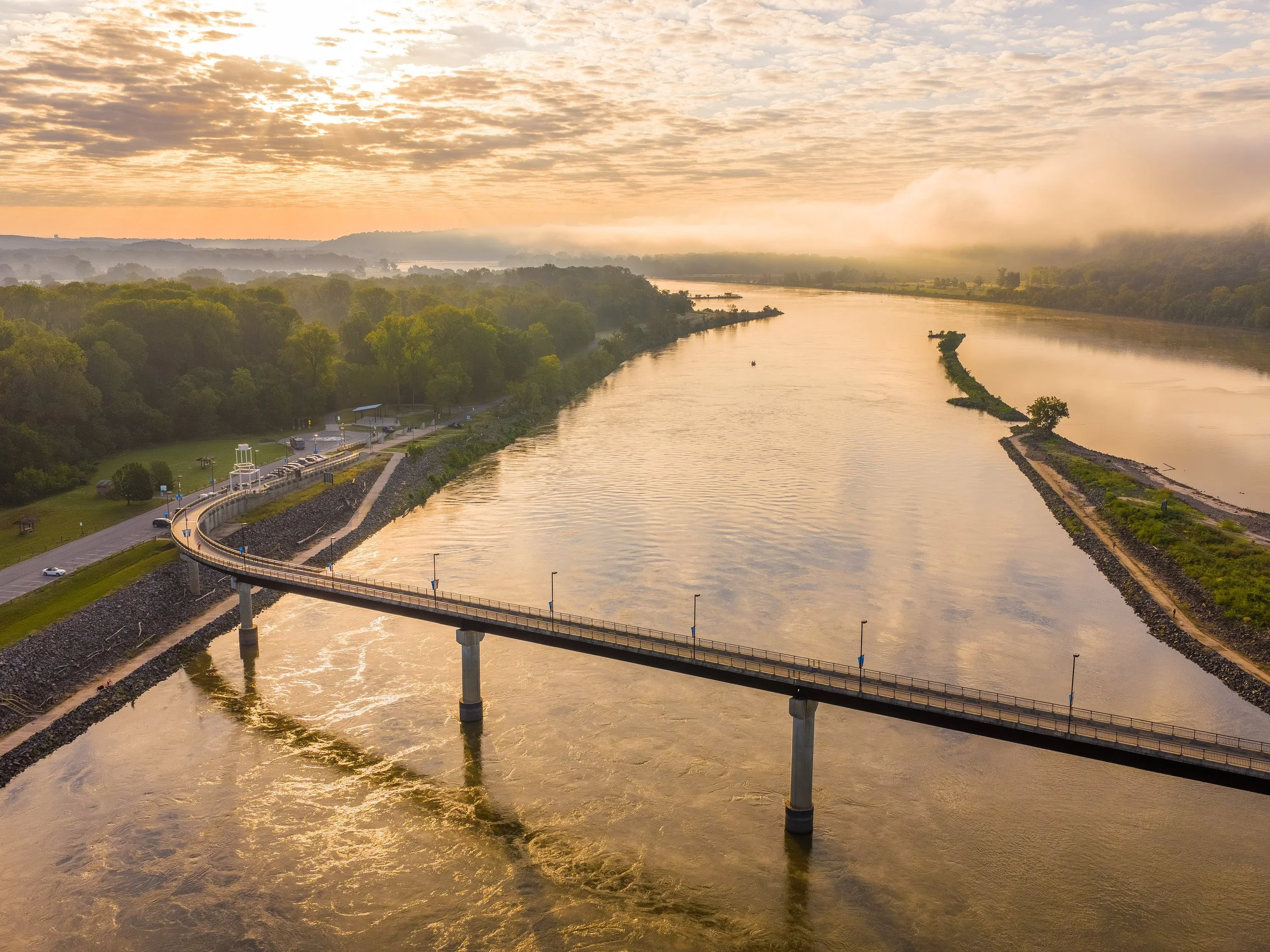 Photo of a river passing under a road bridge at sunset. There are trees along the riverbanks and a cloudy sky with the sun shining through.