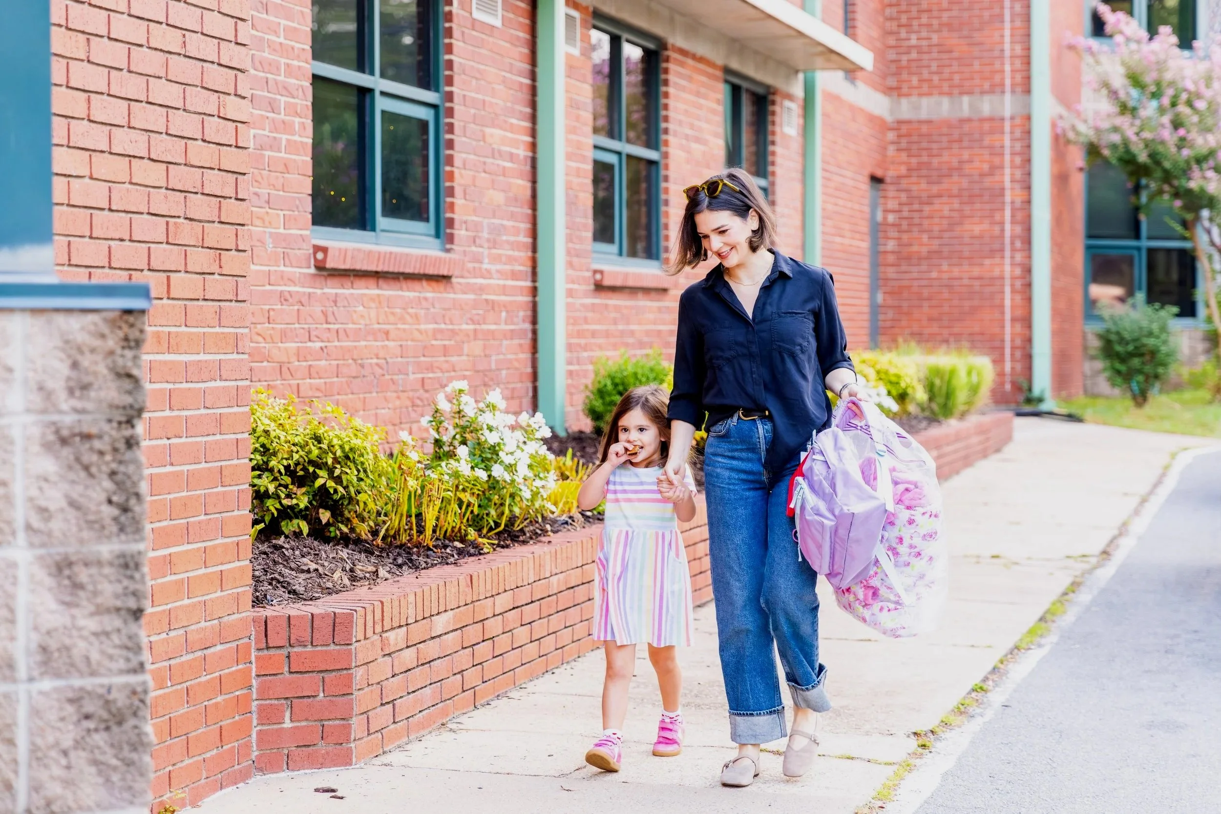 A woman and a young girl walking hand-in-hand on a sidewalk outside a brick building, with the woman carrying a pink backpack and the girl wearing a colorful striped dress.